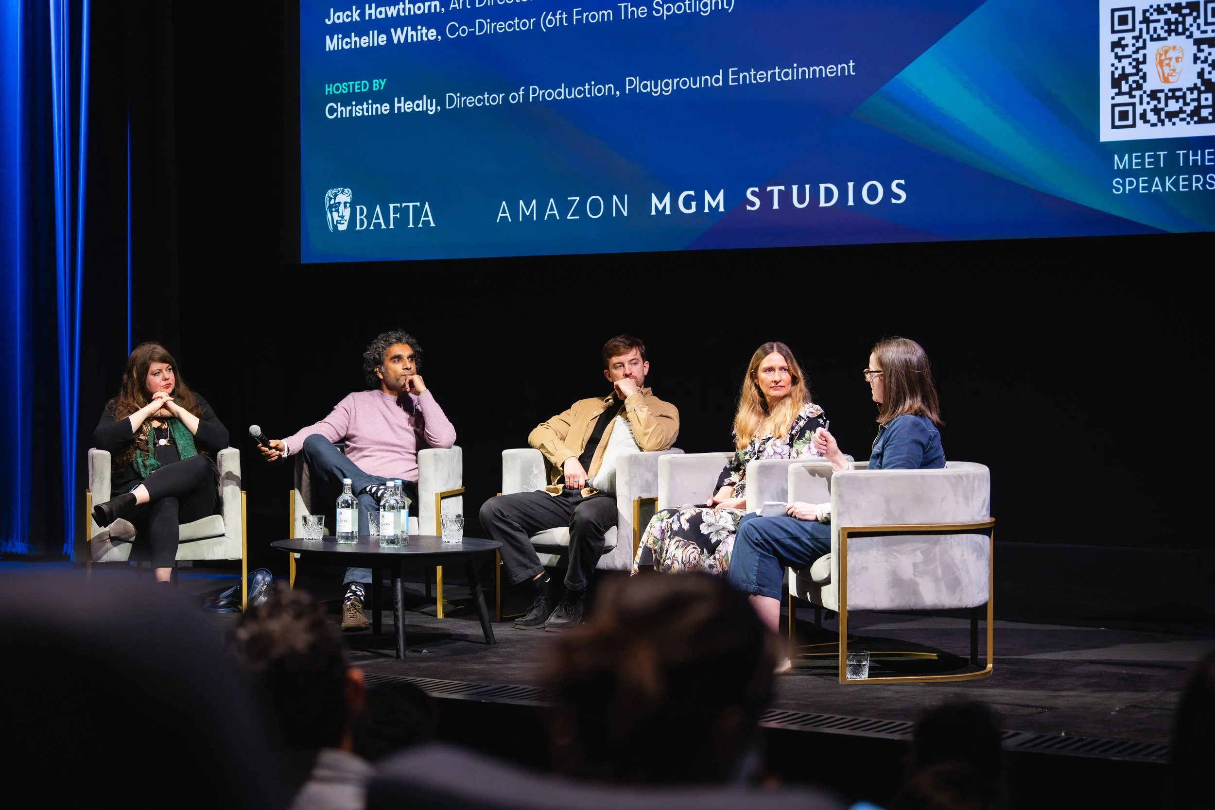 A panel of five people sitting on stage, engaged in conversation, with a large screen behind them displaying text and logos including BAFTA, Amazon, and MGM Studios.