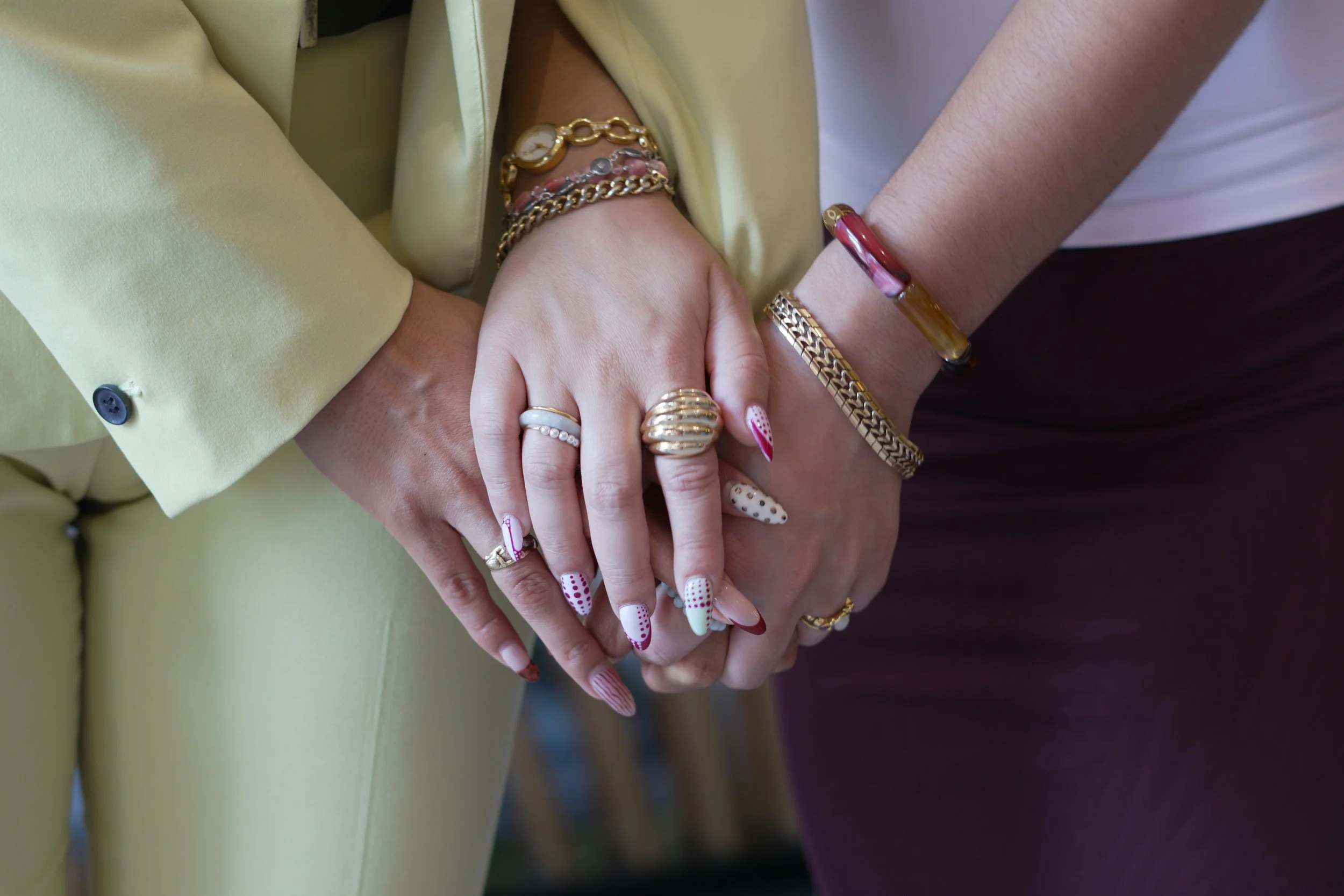 Close-up of two people holding hands, showing their jewelry, including rings and bracelets, with a yellow and a maroon sleeve visible.