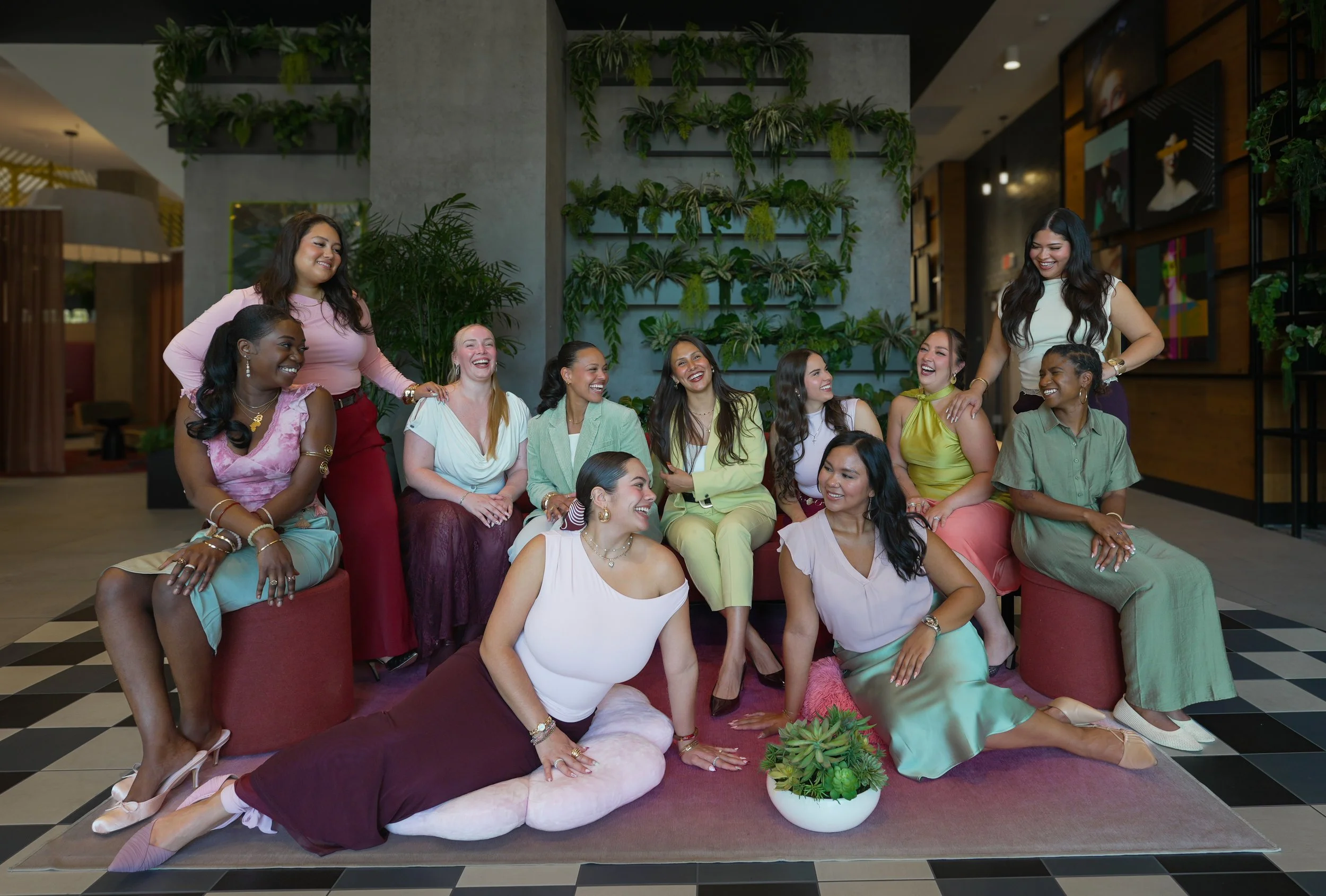 A group of women of diverse ethnicities sitting on a cushioned surface in an indoor space with green wall decor, smiling and enjoying each other's company.