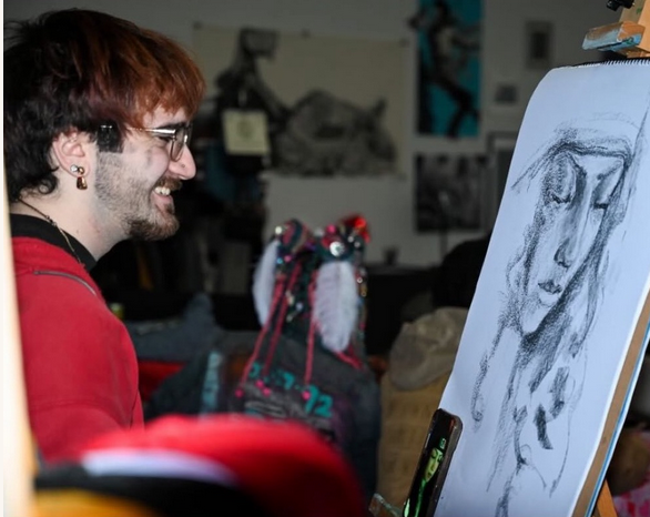 A man with reddish hair, glasses, and a beard smiling while looking at an easel with a charcoal sketch of a woman with long hair and closed eyes.