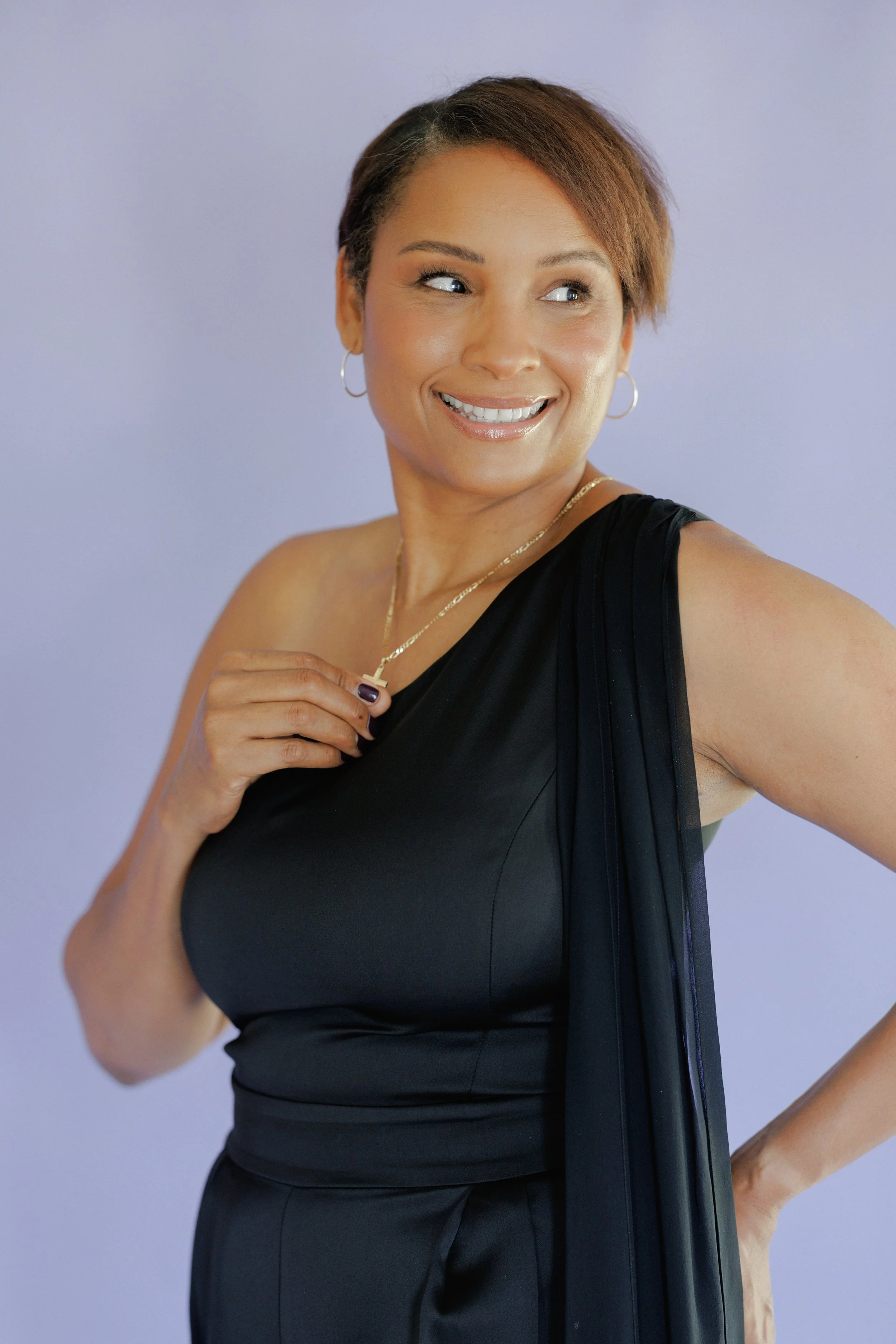 A woman with short brown hair, wearing a black sleeveless top, gold necklace, and hoop earrings, smiling and looking to her right against a plain light purple background.