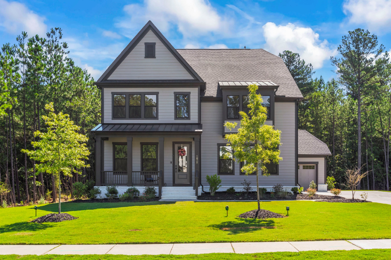 A two-story modern house with gray siding, black window frames, a small front porch, and an attached garage. The house is surrounded by a green lawn with small trees and shrubs, set against a background of tall trees and a blue sky with clouds.