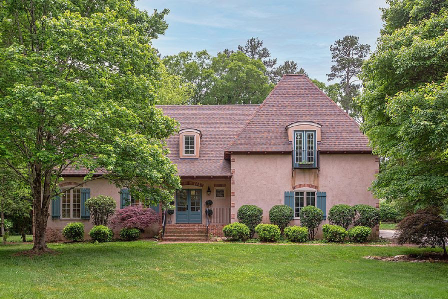 A pink stucco house with blue shutters surrounded by green trees and a well-maintained lawn.