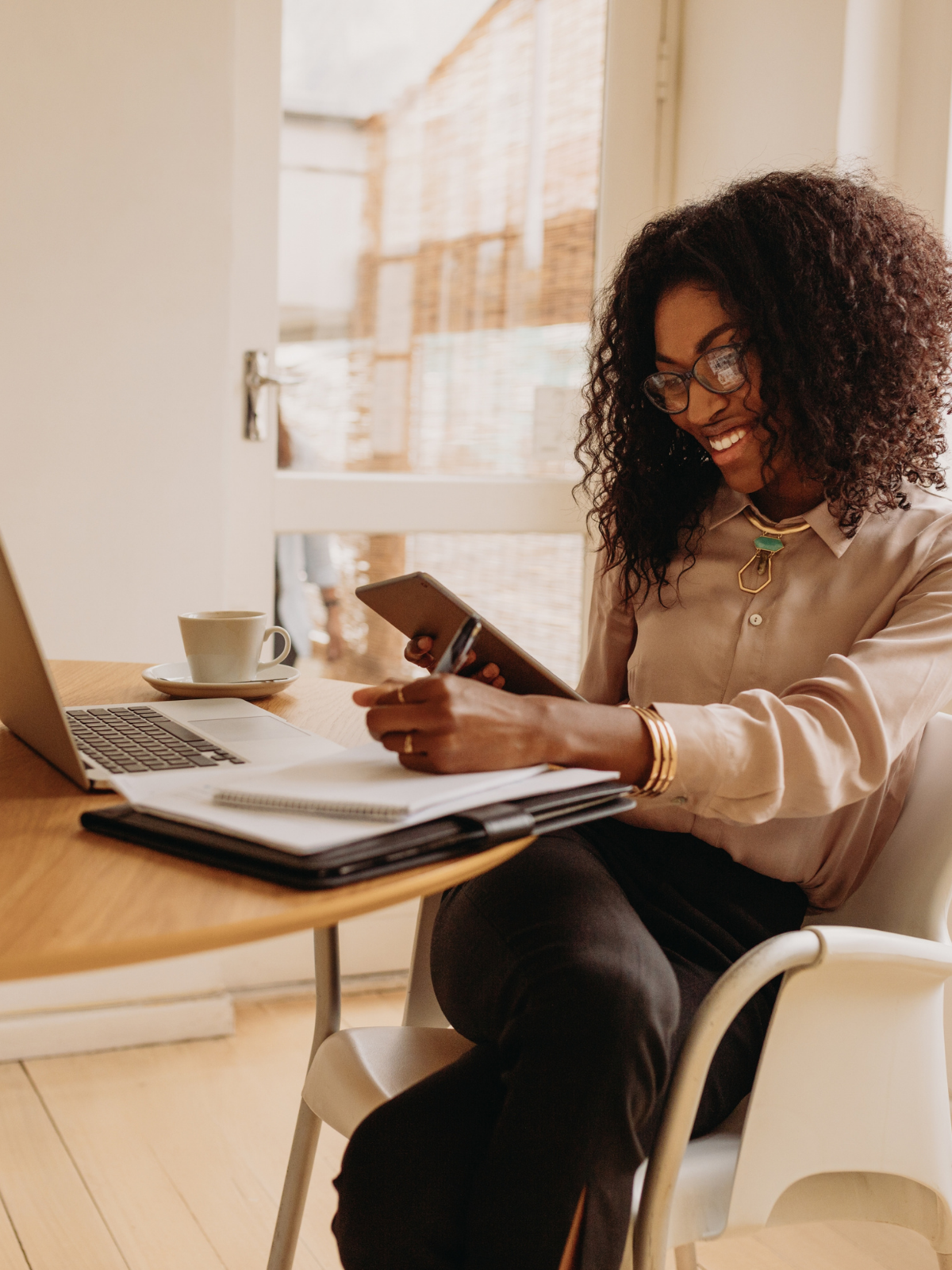 Woman with curly hair and glasses sitting at a desk, smiling, holding a smartphone. On the desk are a laptop, a cup and saucer, notebooks, and pens. She is wearing a beige blouse, layered jewelry, and black pants, in a bright room with a window behind her.