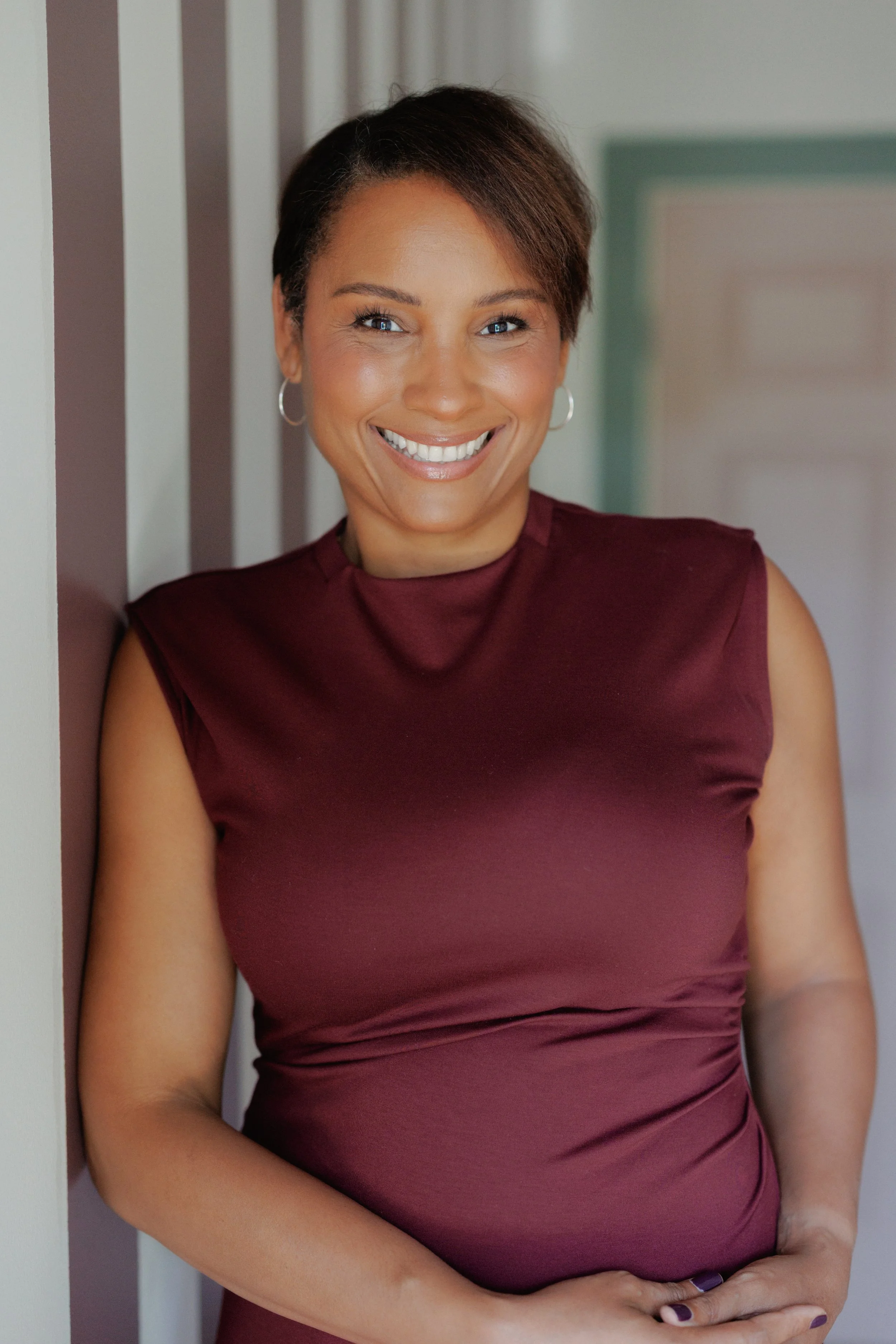 A woman with short brown hair and hoop earrings smiling while leaning against a wall in a maroon sleeveless top.