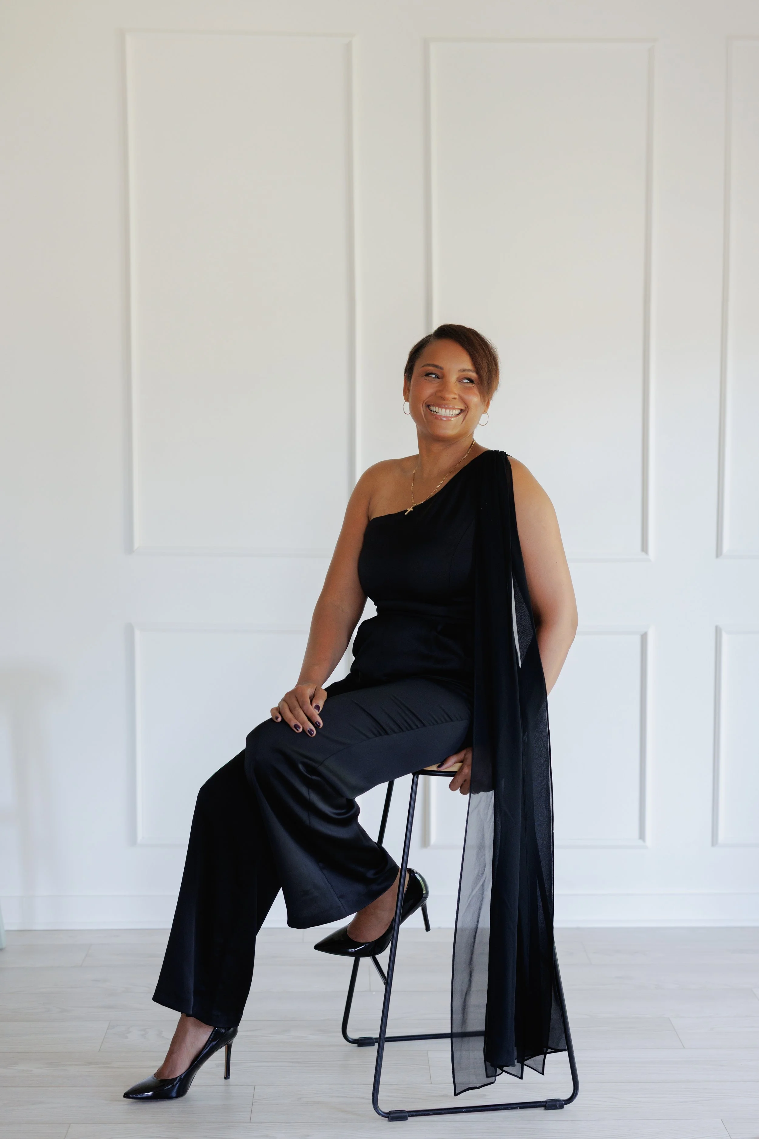 A woman in a sleeveless black dress and high heels sitting on a chair with one leg crossed over the other, smiling. She has short hair and is wearing hoop earrings and a necklace. She is positioned against a white wall with rectangular paneling.