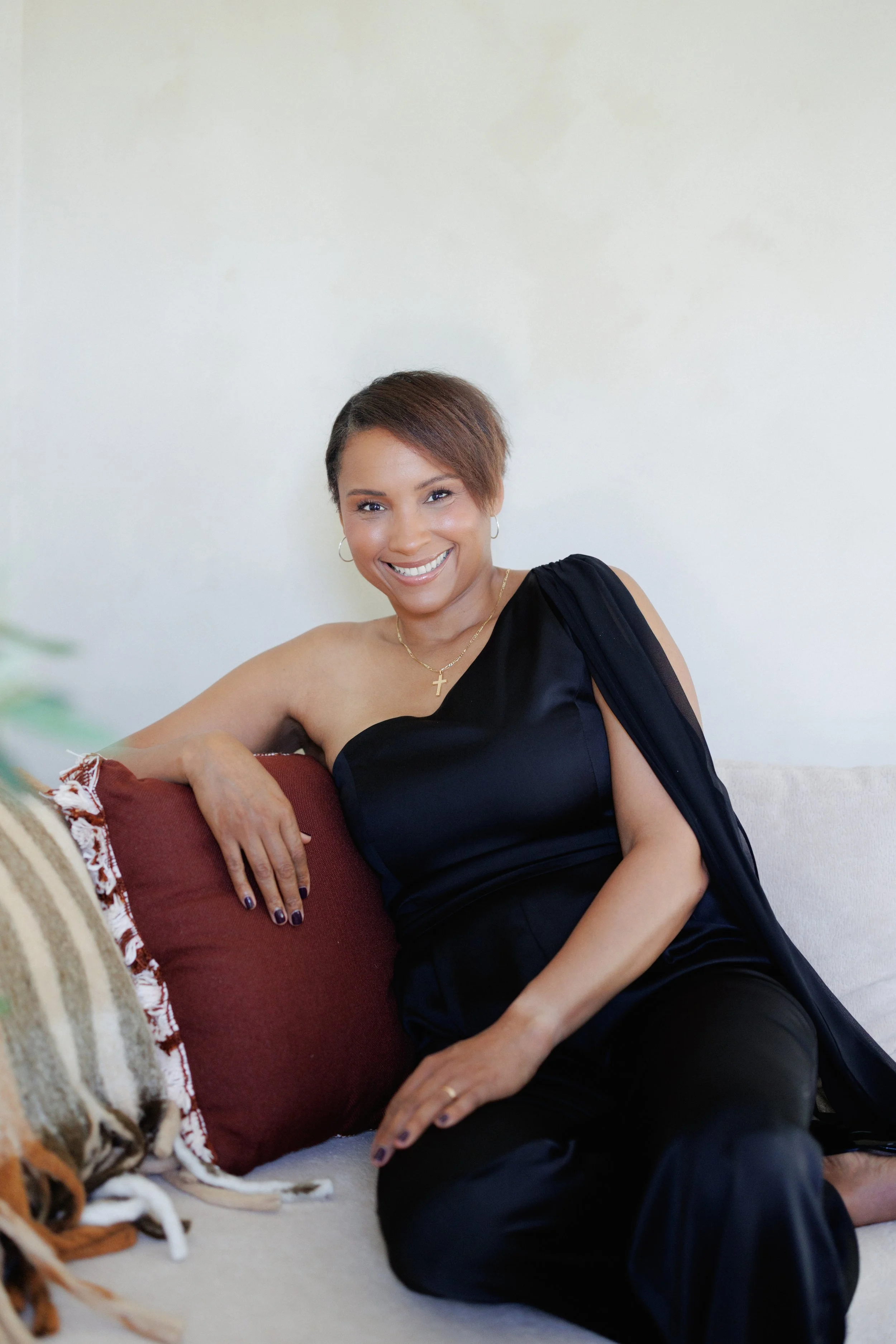 A woman smiling and sitting on the floor, leaning against a sofa with a red cushion. She has short brown hair, earrings, a cross necklace, and is wearing a black dress. There are blankets and a dog partially visible in the foreground.