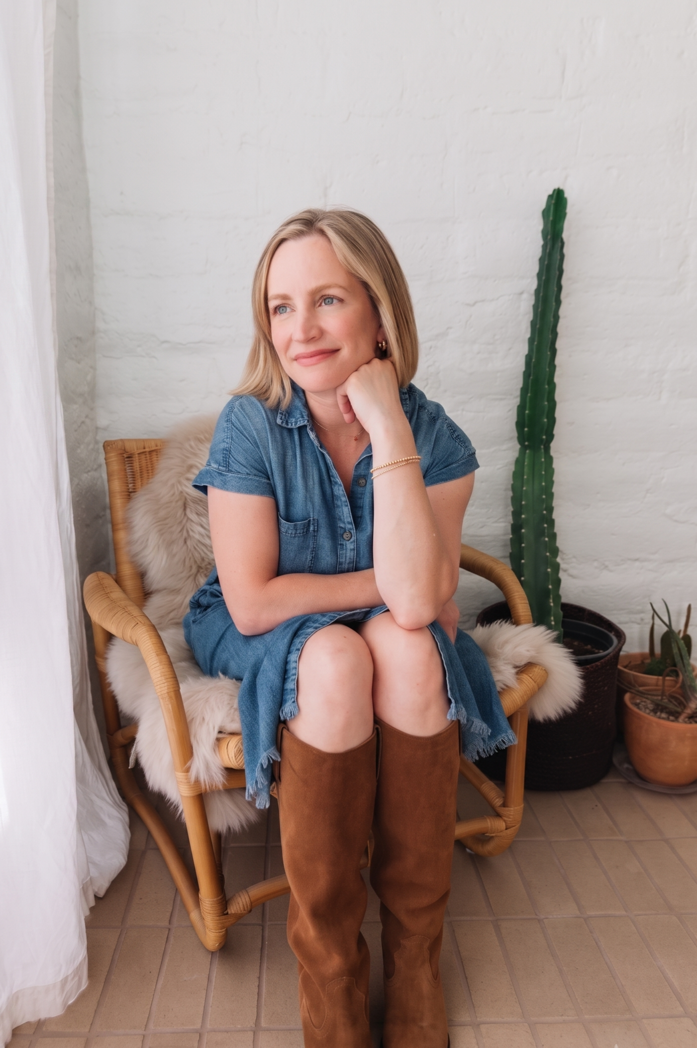 A woman with blonde hair wearing a denim dress, sitting on a rattan chair with a sheepskin throw, in front of a white brick wall with potted cacti, smiling thoughtfully.