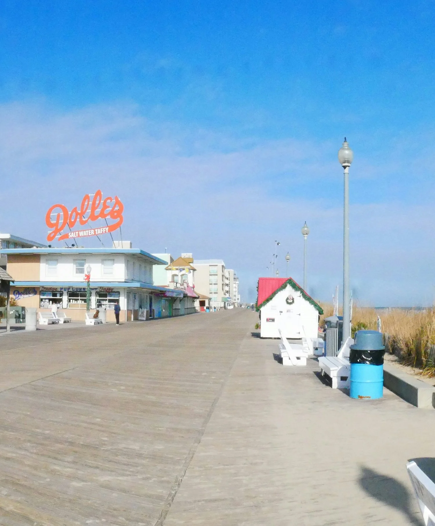 Empty boardwalk with benches, trash cans, lamp posts, and a small hut decorated for Christmas. A large sign for 'Dolle's saltwater taffy' is visible on the building. The sky is clear and blue.