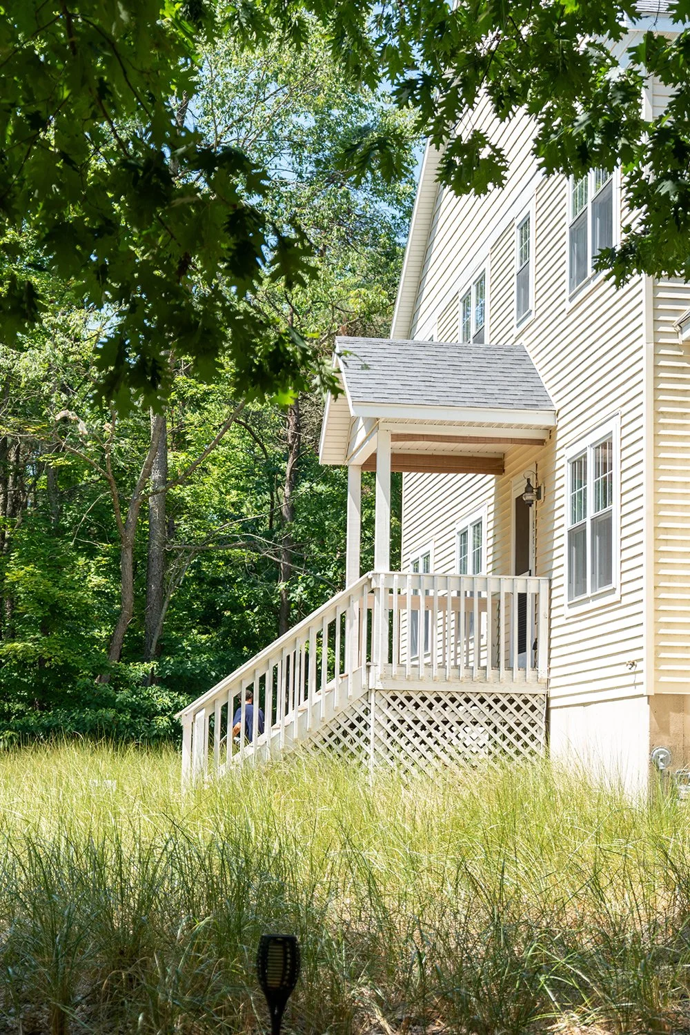 Side view of a beige house with a small porch and stairs, surrounded by trees and grass, with a man walking near the stairs.