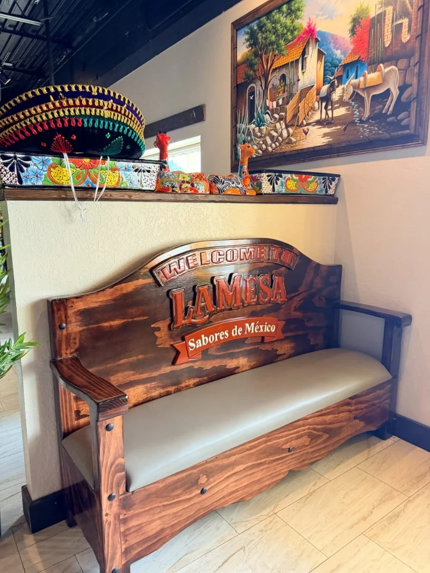Decorative Mexican-themed area with a wooden bench, colorful pottery, a sombrero, and a painting of a village scene with horses.