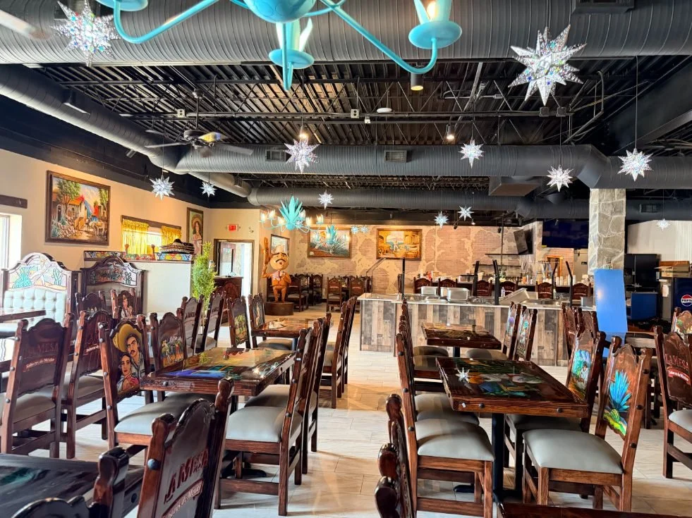 Empty restaurant dining area decorated with hanging snowflake and star-shaped ornaments, themed Mexican artwork, and wooden tables and chairs.