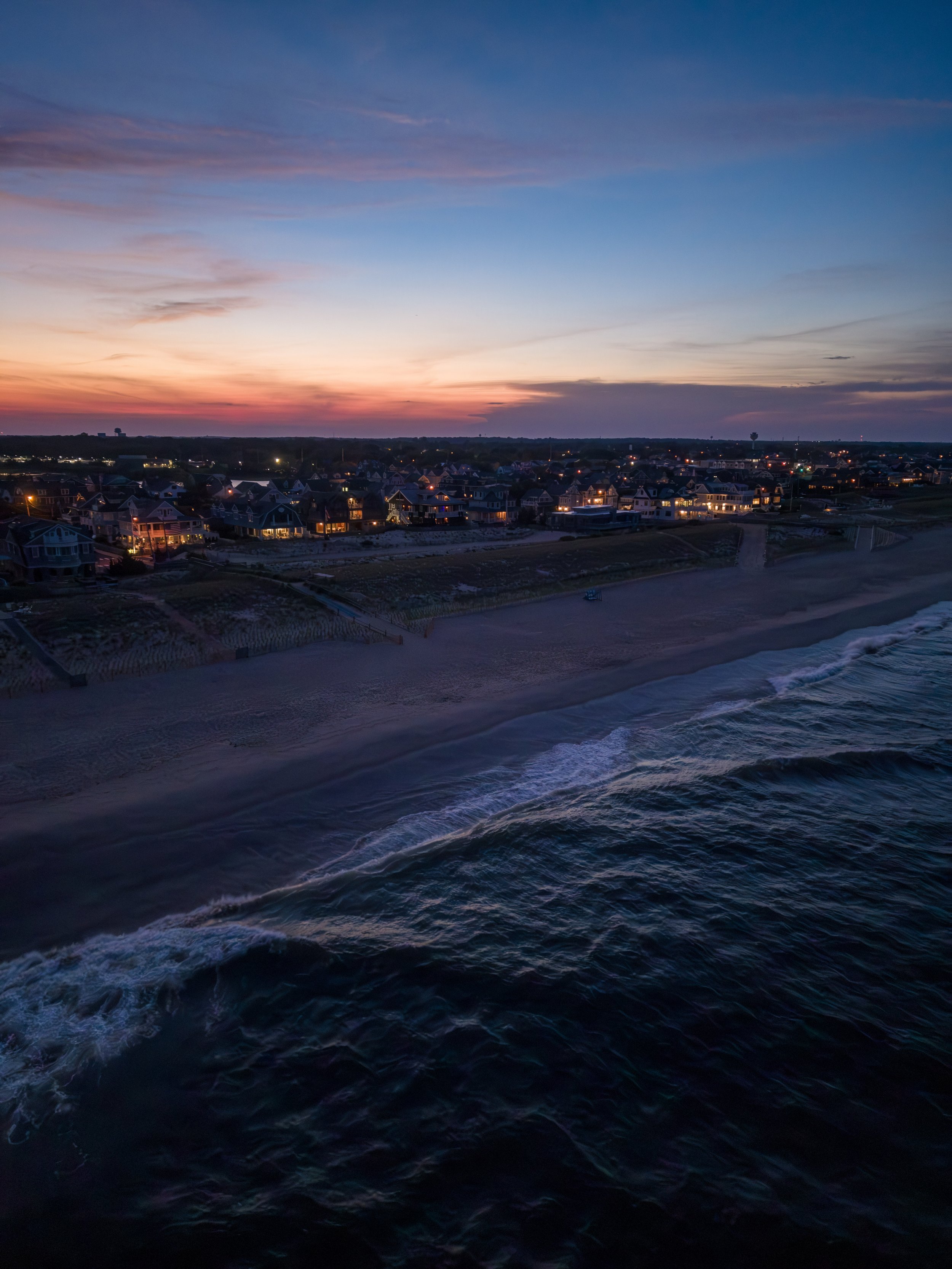 A beachside residential area at dusk with houses facing the ocean, a sandy shoreline, and a colorful sunset sky with clouds.
