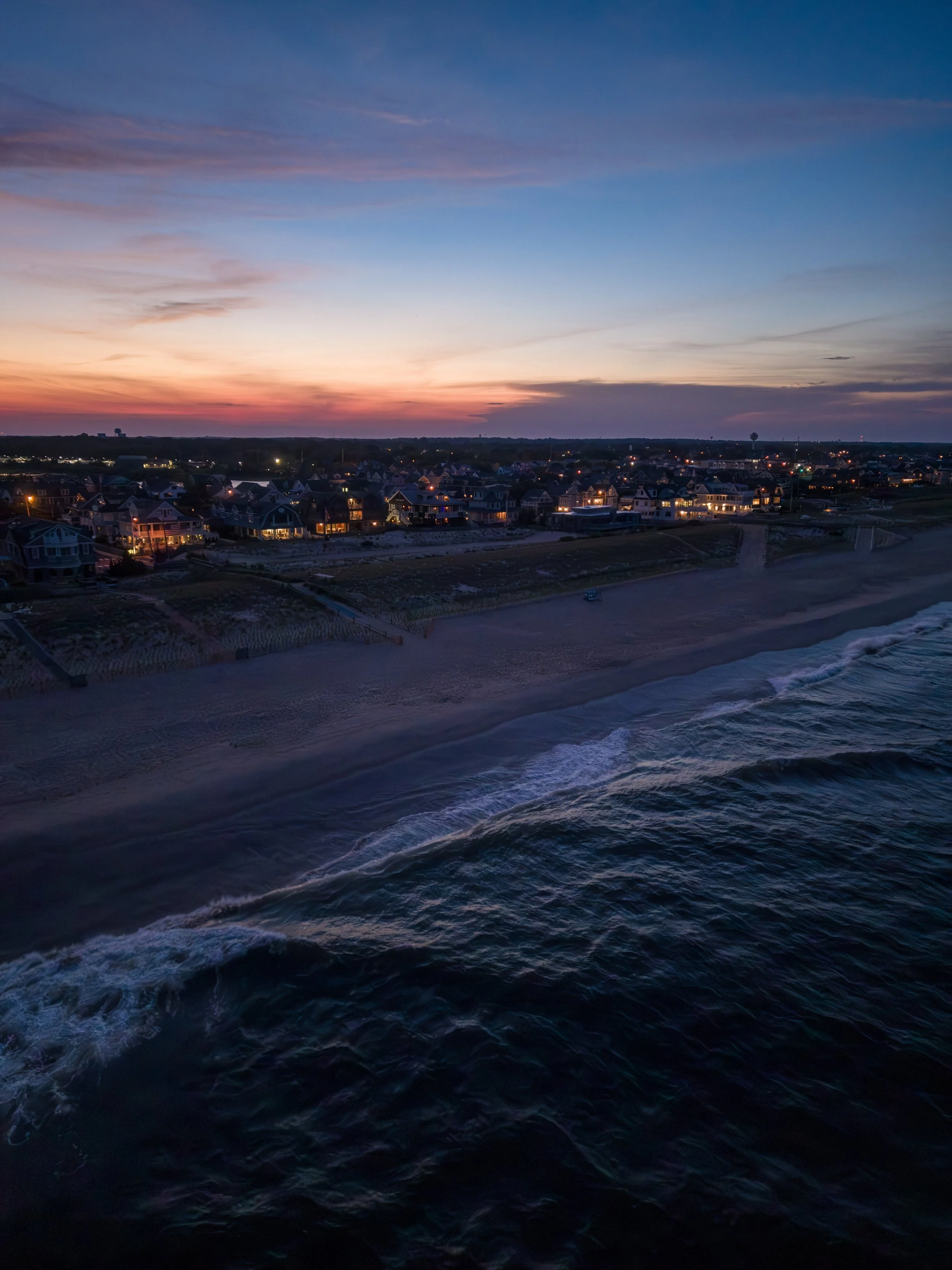 Ocean waves at sunset with a distant coastal town and a colorful sky.
