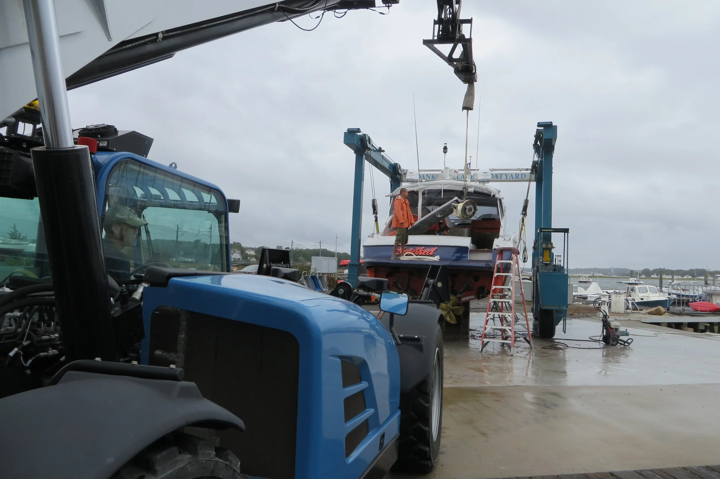 A boat is positioned on a hydraulic lift at a marina, with a worker in an orange jacket inspecting it. The marina has several docked boats, and the sky is overcast.