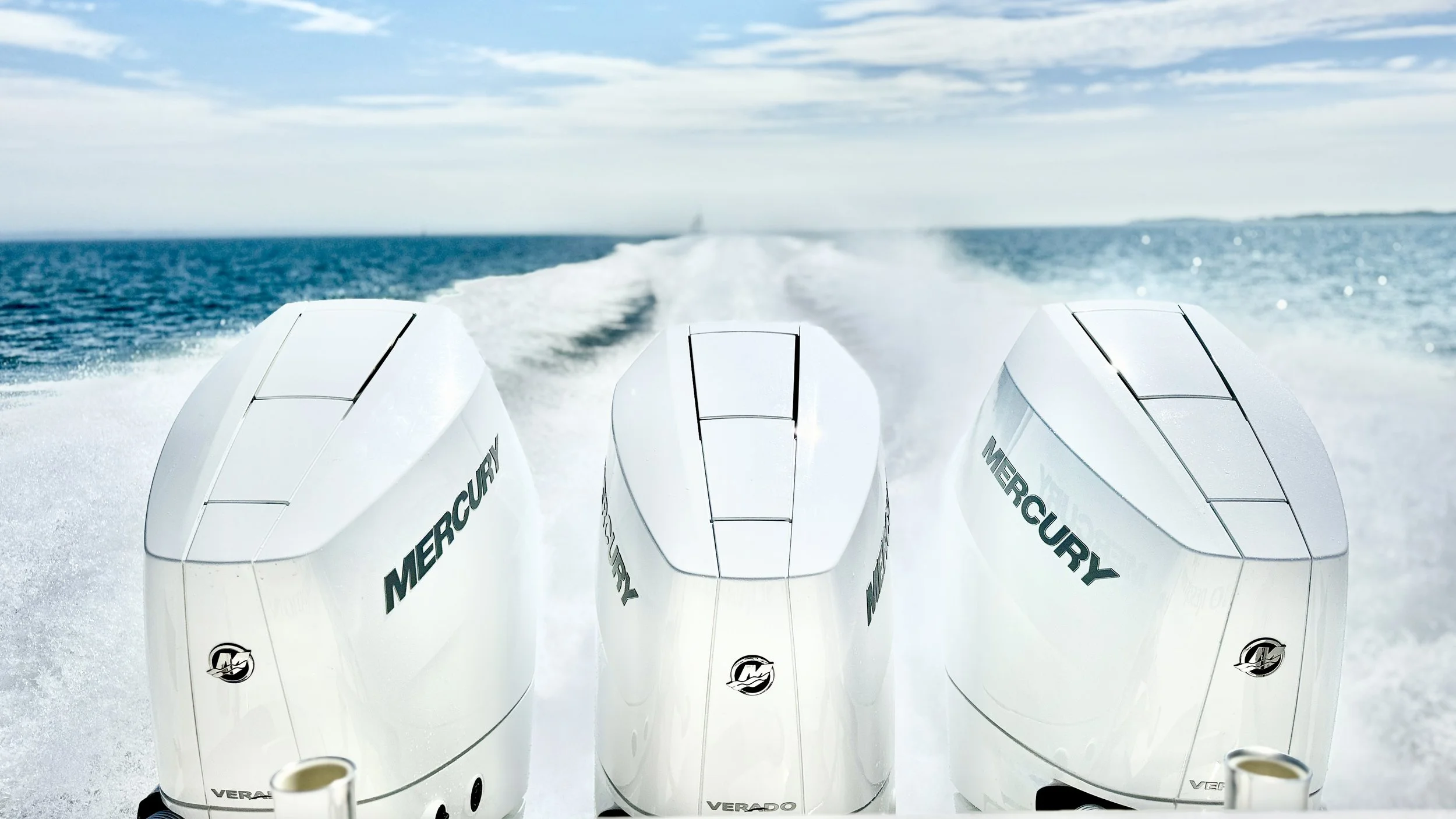 The photo shows the back of a boat moving across the water with three Mercury outboard engines attached to its stern, creating a wake behind it.