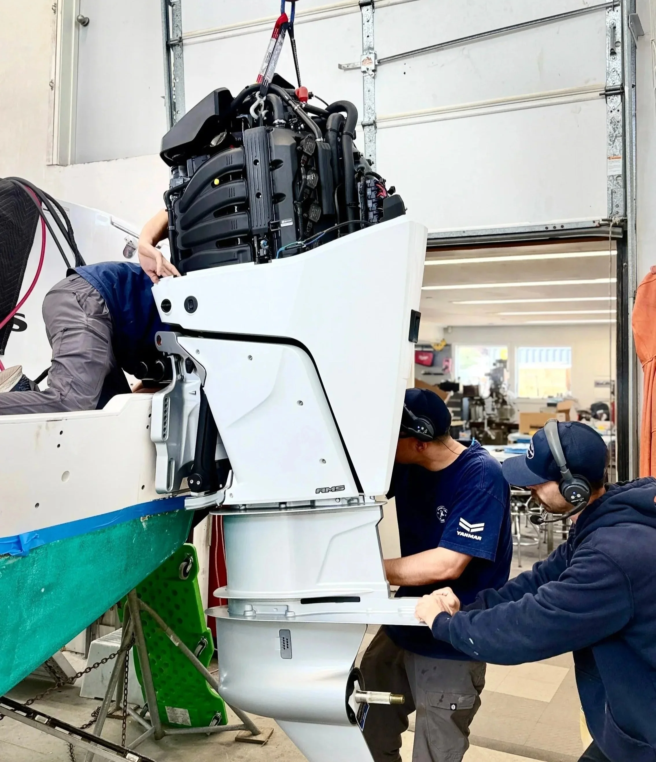 Two technicians are working on a large internal component of a spacecraft or advanced machinery in a workshop. One is wearing a dark blue shirt with a logo on the sleeve and a cap, the other is wearing black. They are using tools on the equipment, which is mounted vertically and appears to be in an assembly or maintenance process. The background includes workshop tools, equipment, and a green container.