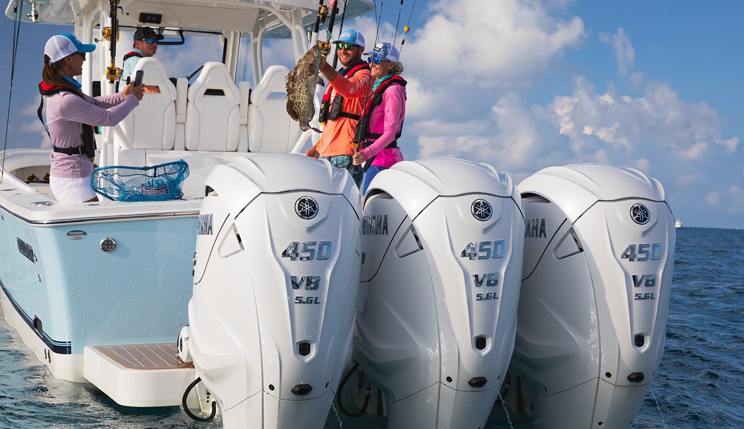 A group of four people on a boat, with three of them holding a large fish they caught. The boat has three Yamaha 450 V8 outboard motors and is out on the water with clouds in the sky.