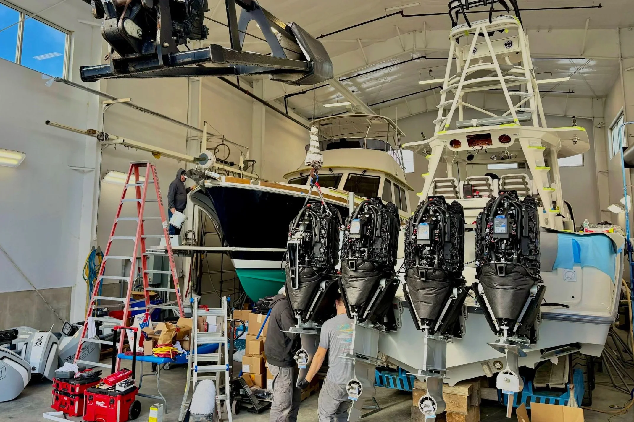 Workers assembling a large yacht inside a workshop with tools, equipment, and engines visible, with parts suspended from the ceiling.