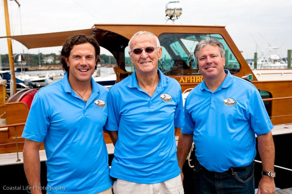 Three men wearing matching blue polo shirts standing in front of a wooden boat at a marina.