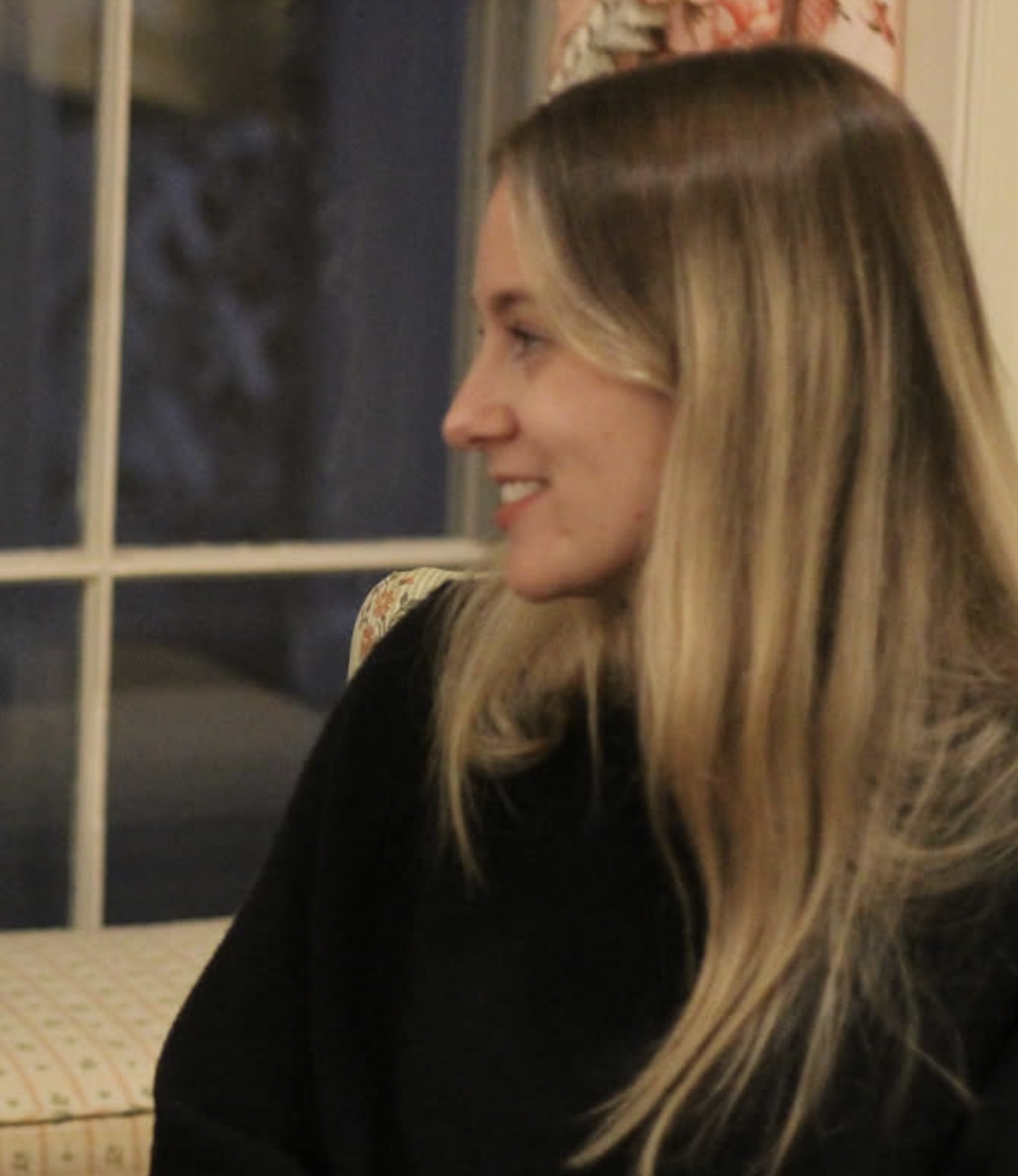 A woman with long blonde hair smiling and looking to the side, sitting indoors near a window.