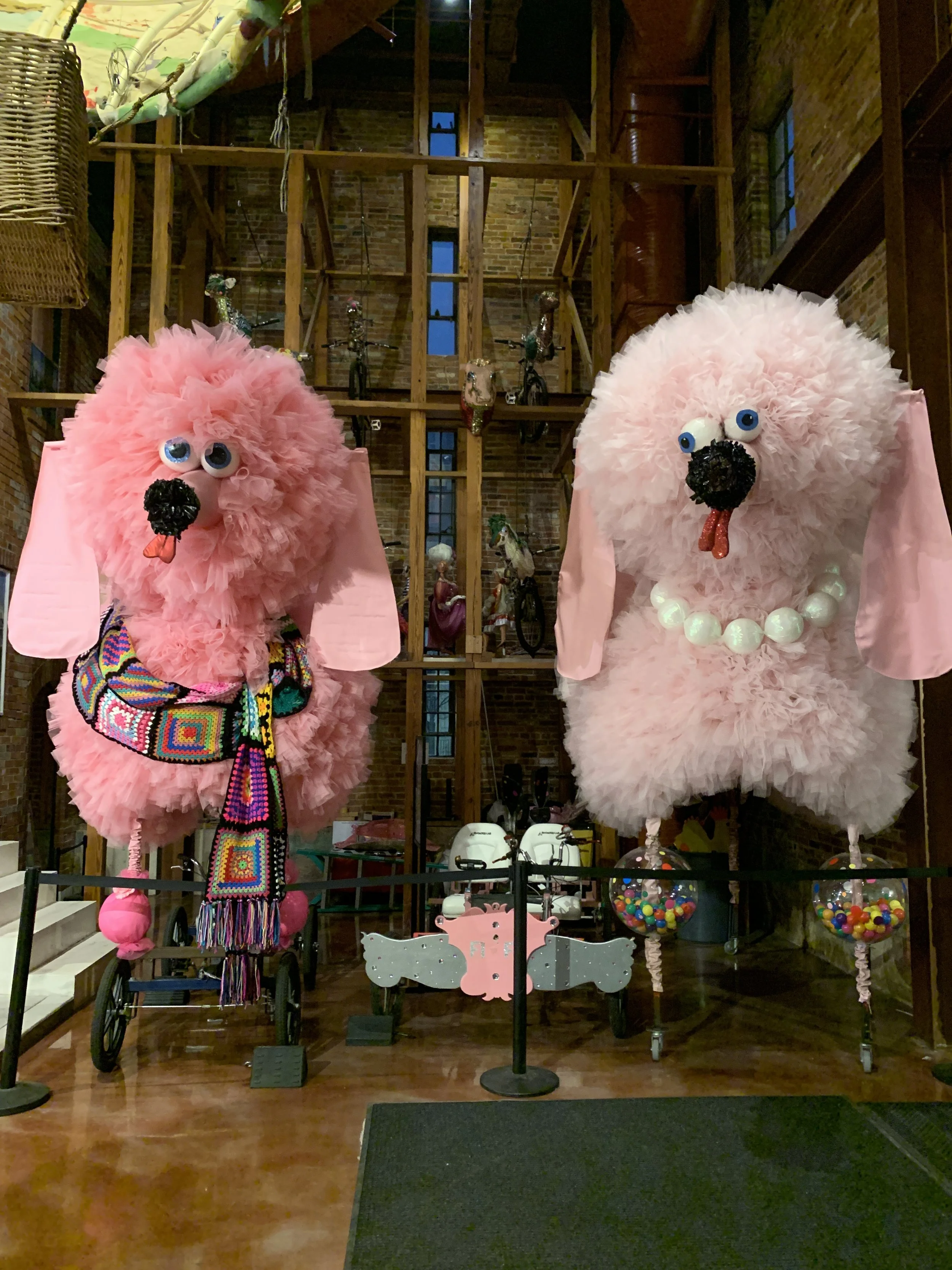 Decorative pink poodles with large fluffy fur, googly eyes, black noses, and red tongues on bottom, with colorful accessories and beads, displayed inside a brick-and-wood interior with shelves, bicycles, and toys.