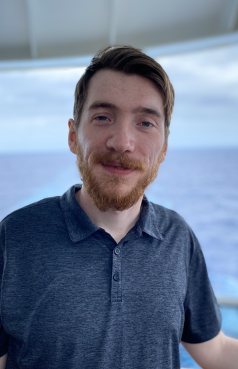 A young man with light skin, brown hair, and a beard, smiling while standing on a boat with the ocean in the background.