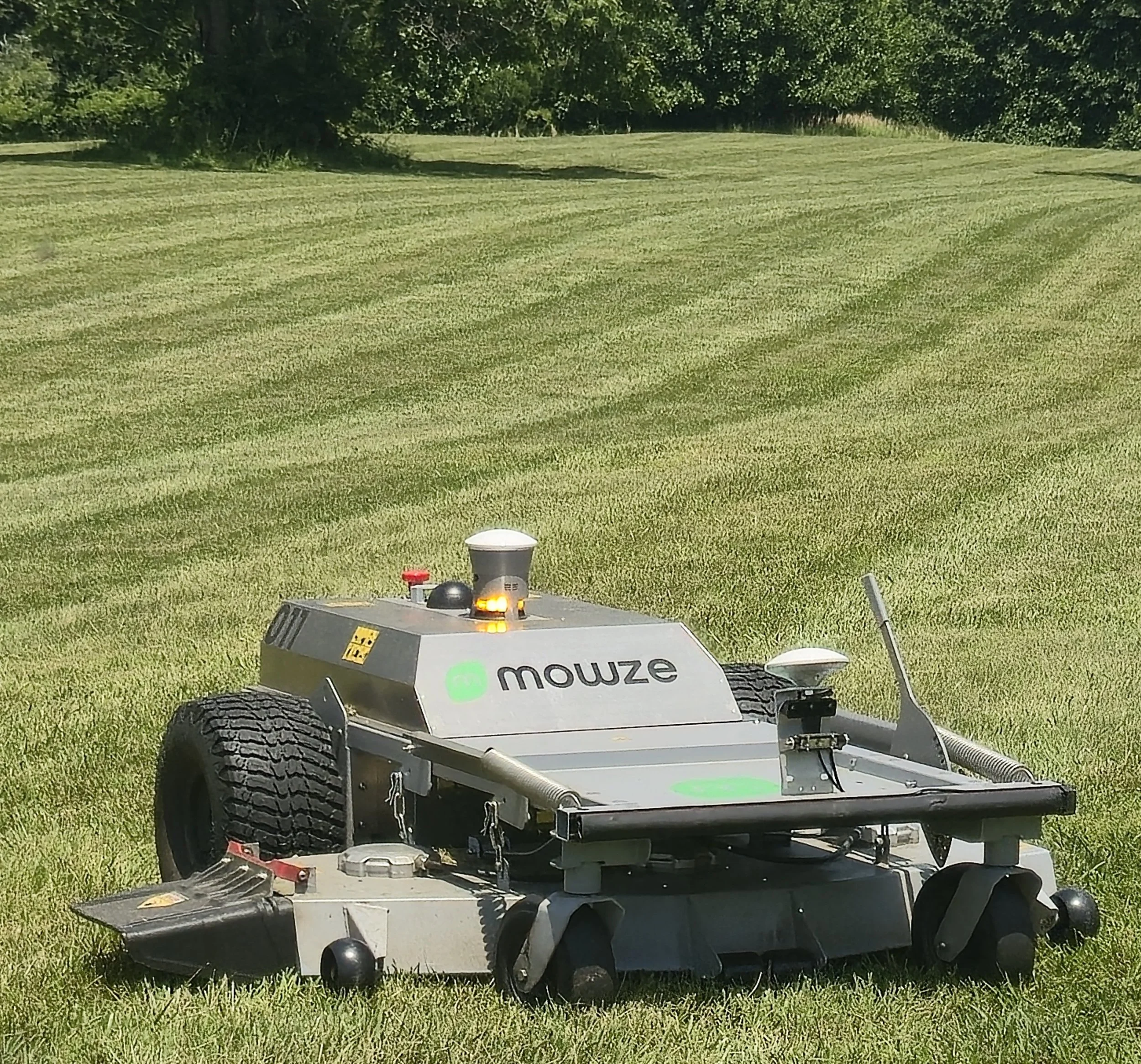 The MOWZE M60 all electric robotic mower on a grassy field, with trees in the background.