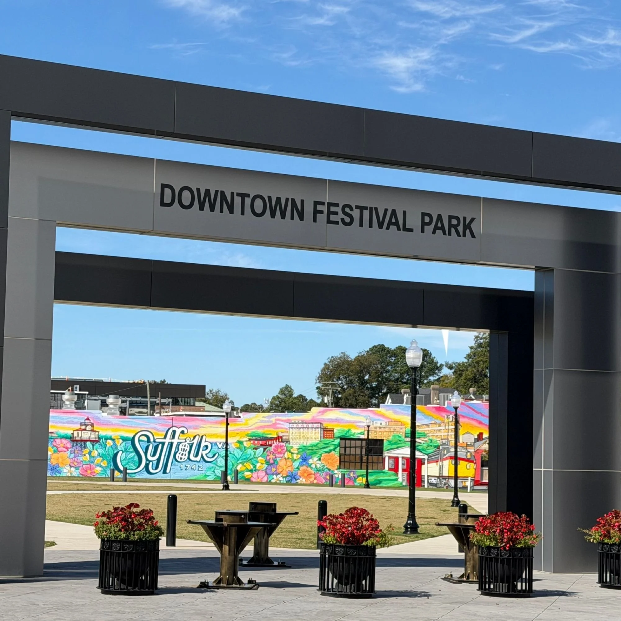 View of an entrance area in Suffolk, Virginia featuring potted red flowers, tables, and lampposts, with a colorful mural and blue sky in the background.