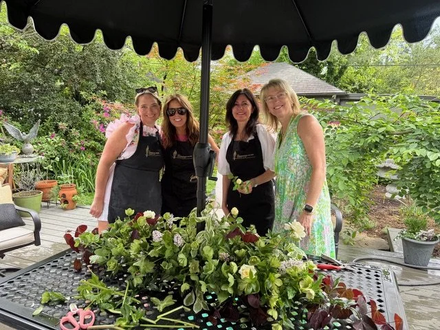 Four women stand under a black patio umbrella, smiling behind a table with a large floral arrangement, in a garden setting.