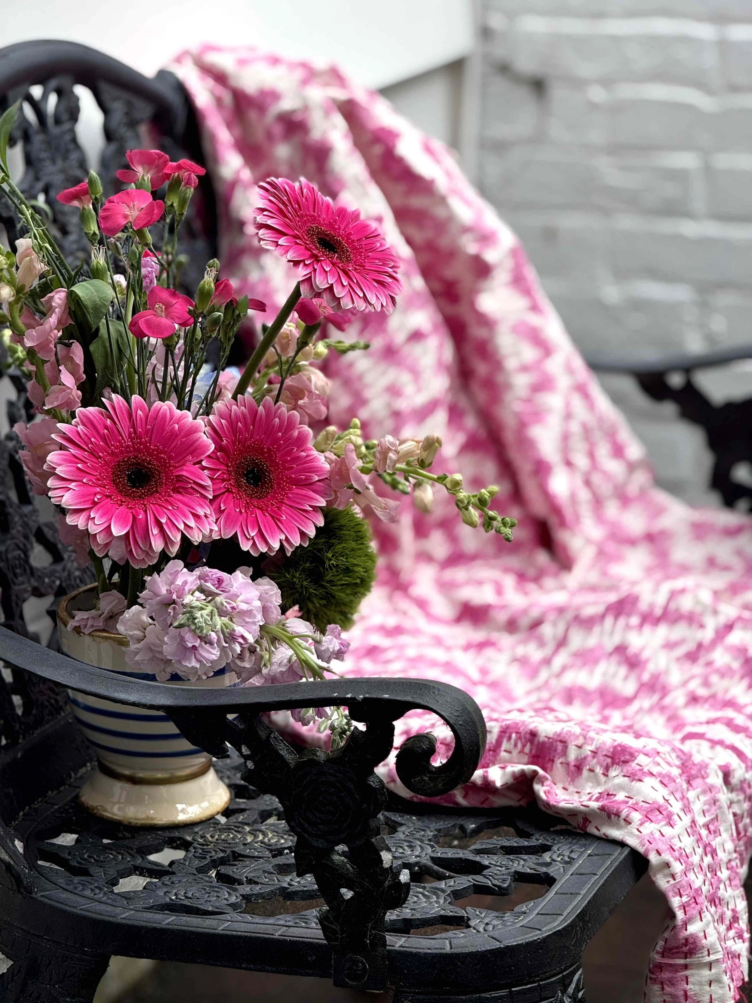 A bouquet of pink flowers, including gerbera daisies and other blossoms, in a striped ceramic pot, placed on a decorative black metal table, with pink and white patterned fabric in the background.