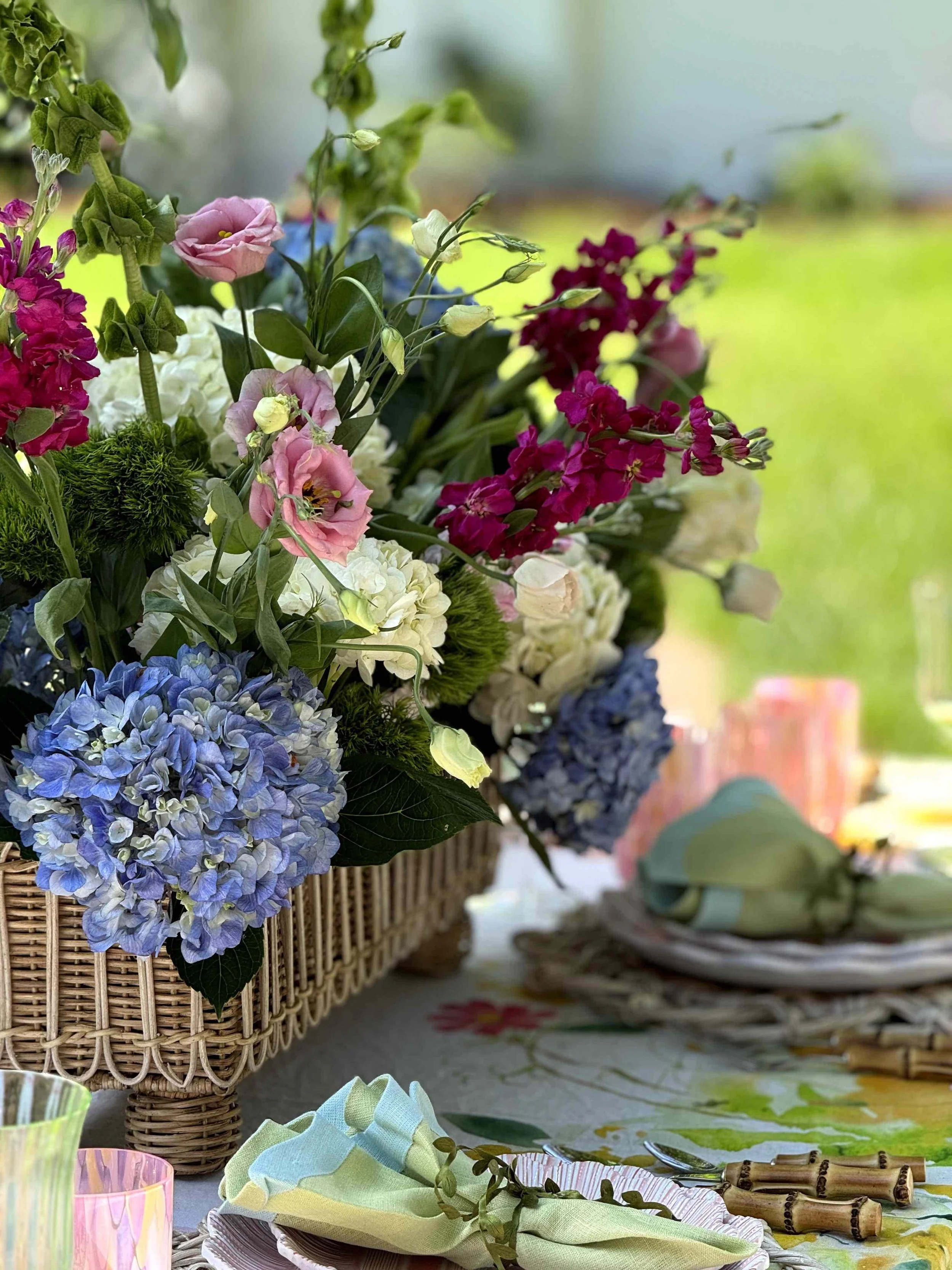 Colorful flower arrangement with hydrangeas, lisianthus, and other flowers in a wicker basket on a table, with pastel-colored glasses, napkins, bamboo utensils, and a floral tablecloth in an outdoor setting.