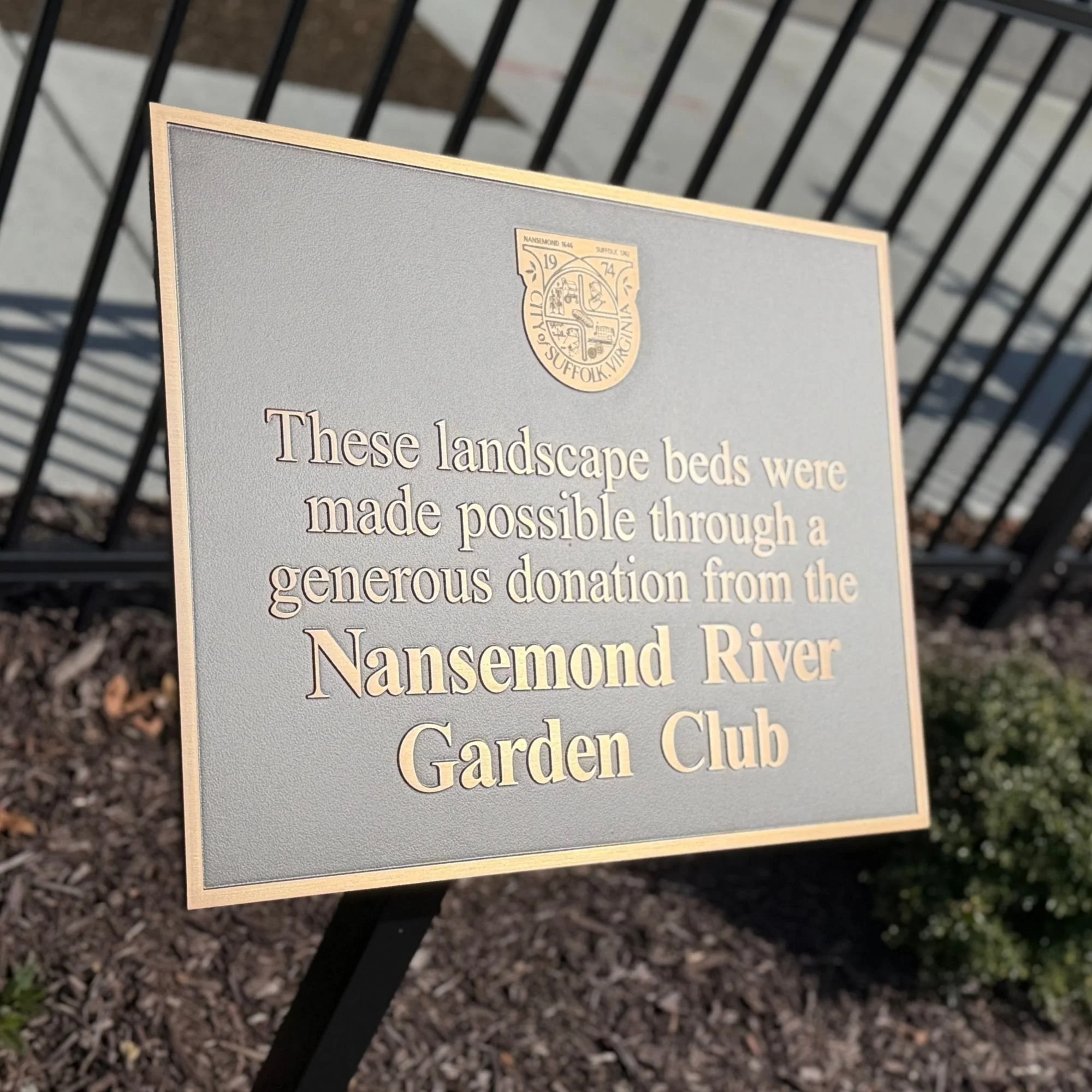 Close-up of a sign with text about landscape beds and a town crest, with a black metal fence and gravel ground in the background.