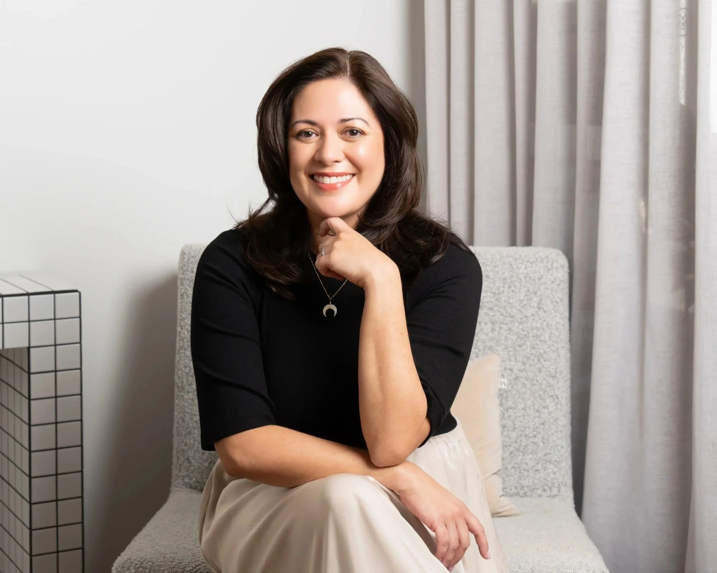 A woman with shoulder-length dark brown hair smiling and resting her chin on her hand, sitting on a light-colored chair with a cream-colored curtain behind her and a white wall to her left.