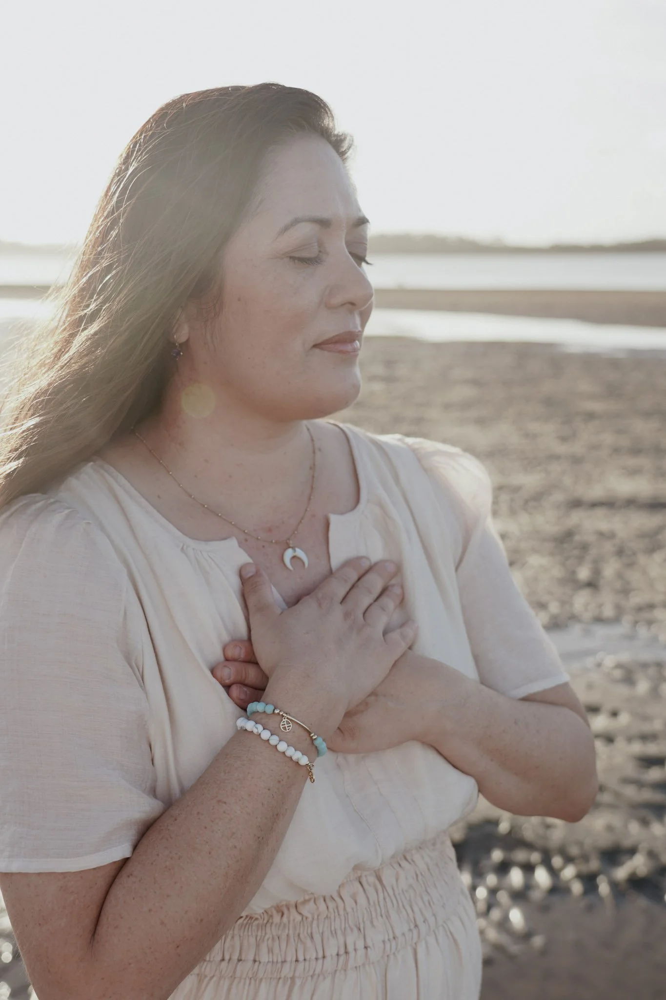 A woman with long hair, closed eyes, and a serene expression standing outdoors on a beach at sunset, with her hand on her chest.