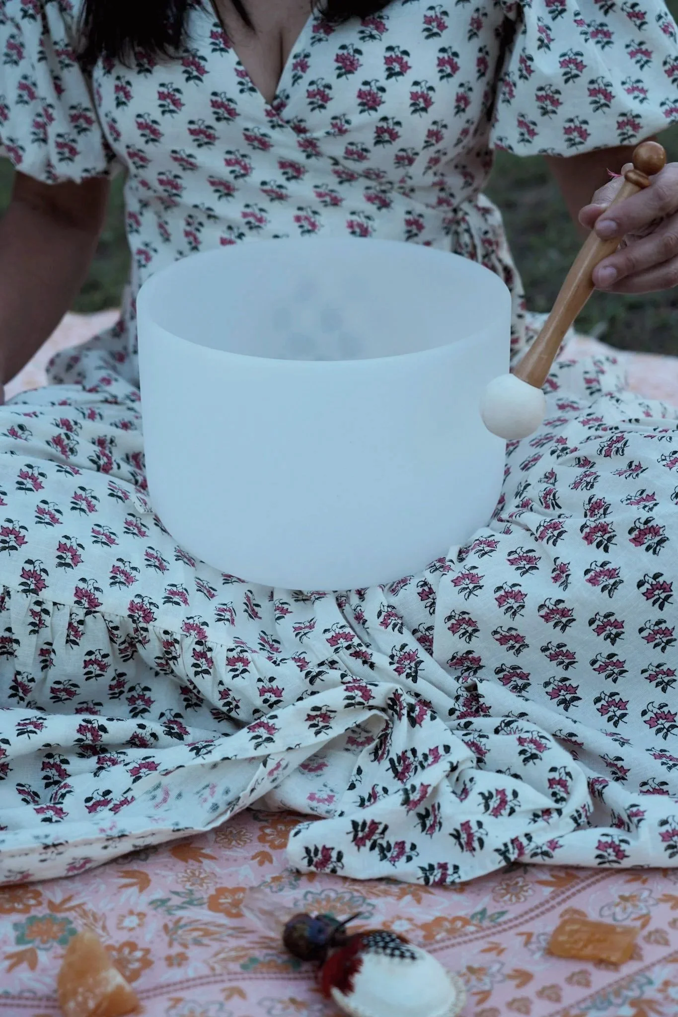 Person sitting on a blanket wearing a floral dress, holding a singing bowl with a wooden mallet, with crystal and wellness items in front.