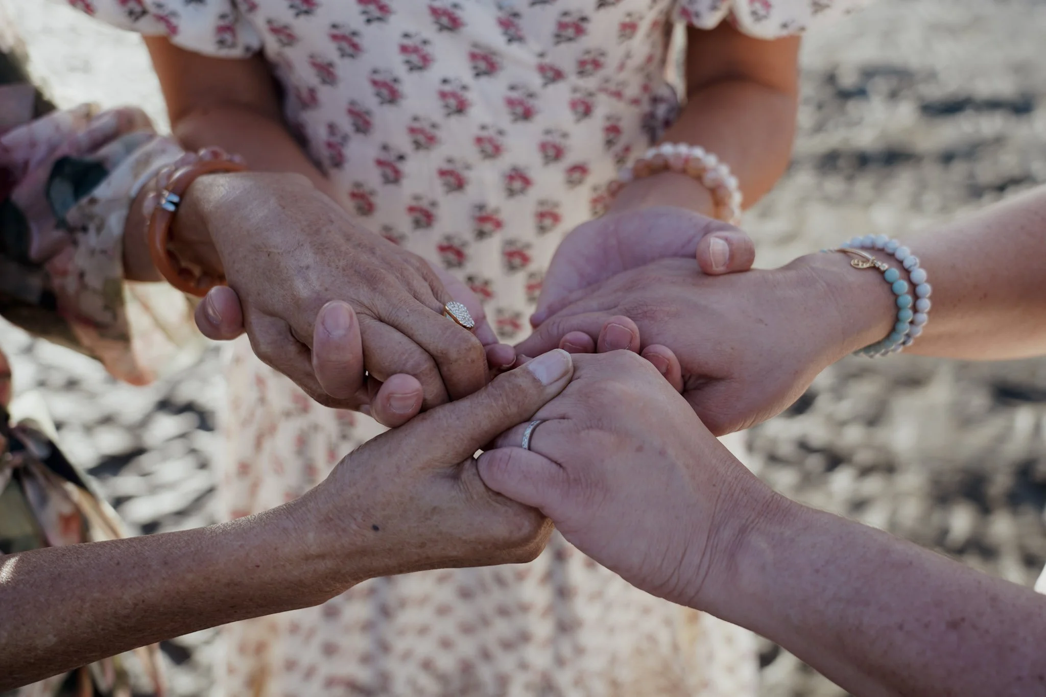 Multiple hands of different ages and skin tones joined together in a circle, with some wearing jewelry, on a sandy background.
