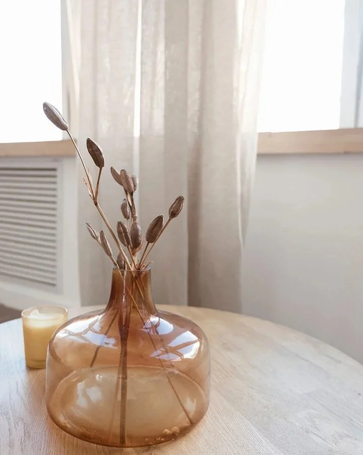 Brown glass vase with dried bunny tail grass on a wooden table, with a small white candle to the left and curtains in the background.