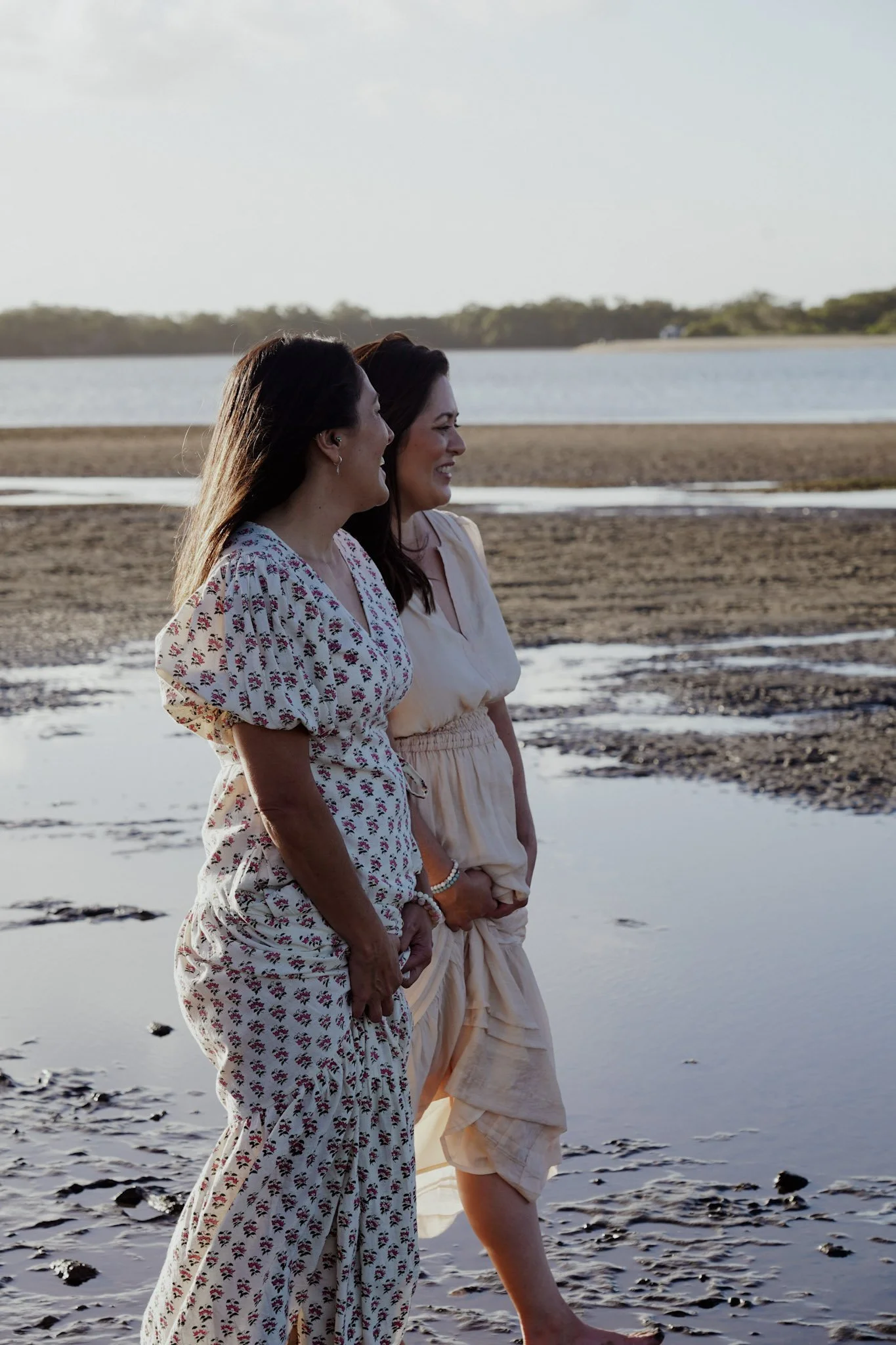 Two women with long dark hair standing on a sandy and rocky shoreline near water, smiling and looking to the right during the daytime.