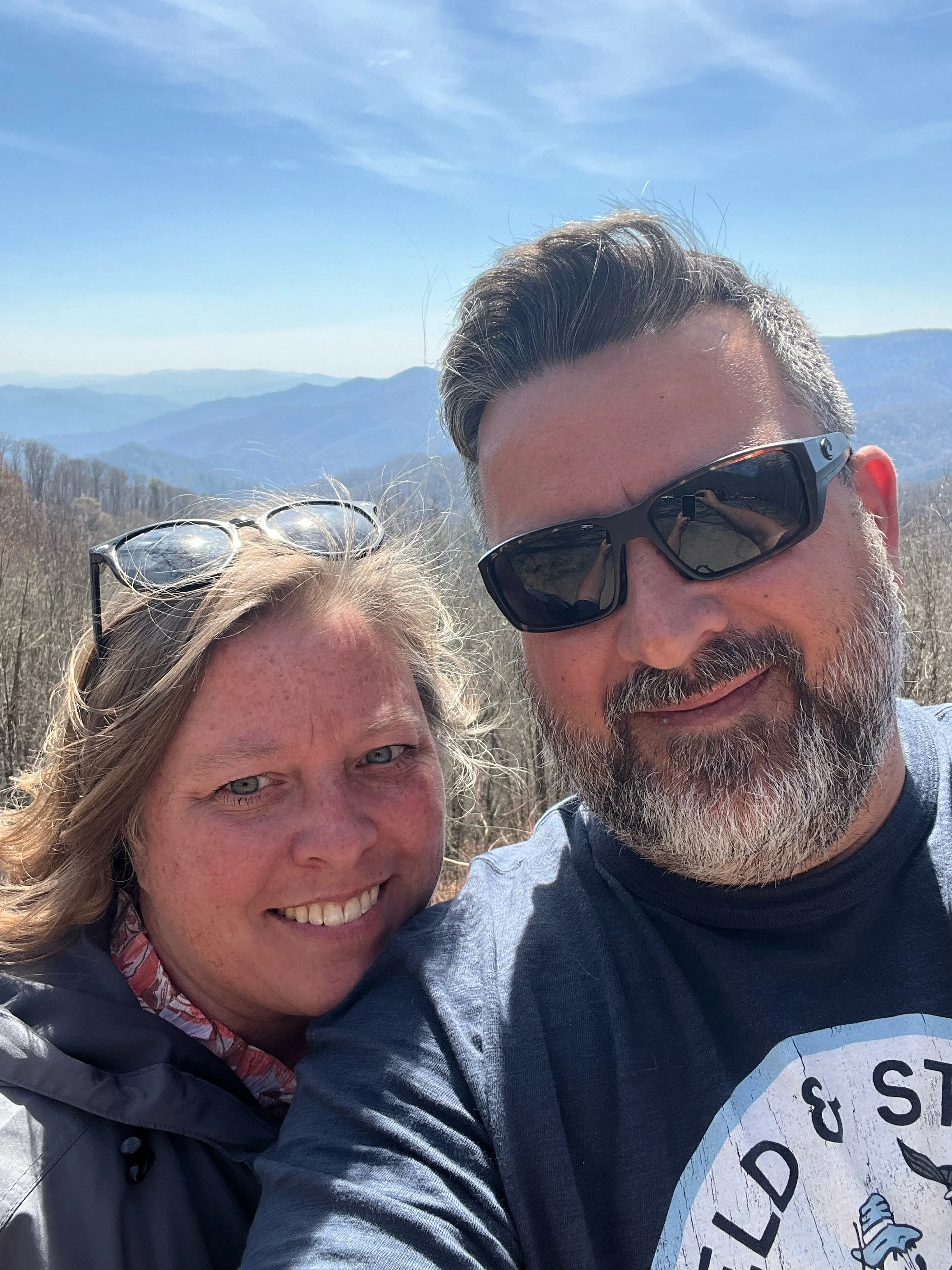 A smiling woman and a man with sunglasses taking a selfie outdoors with mountains and a blue sky in the background.