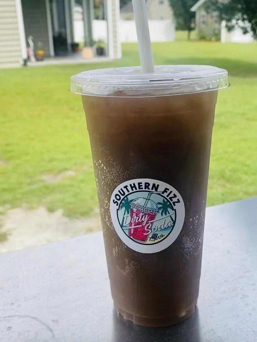 A plastic cup filled with iced coffee or similar beverage, with a clear lid and straw, placed on a white surface, outdoors with a lawn, trees, and a house in the background.