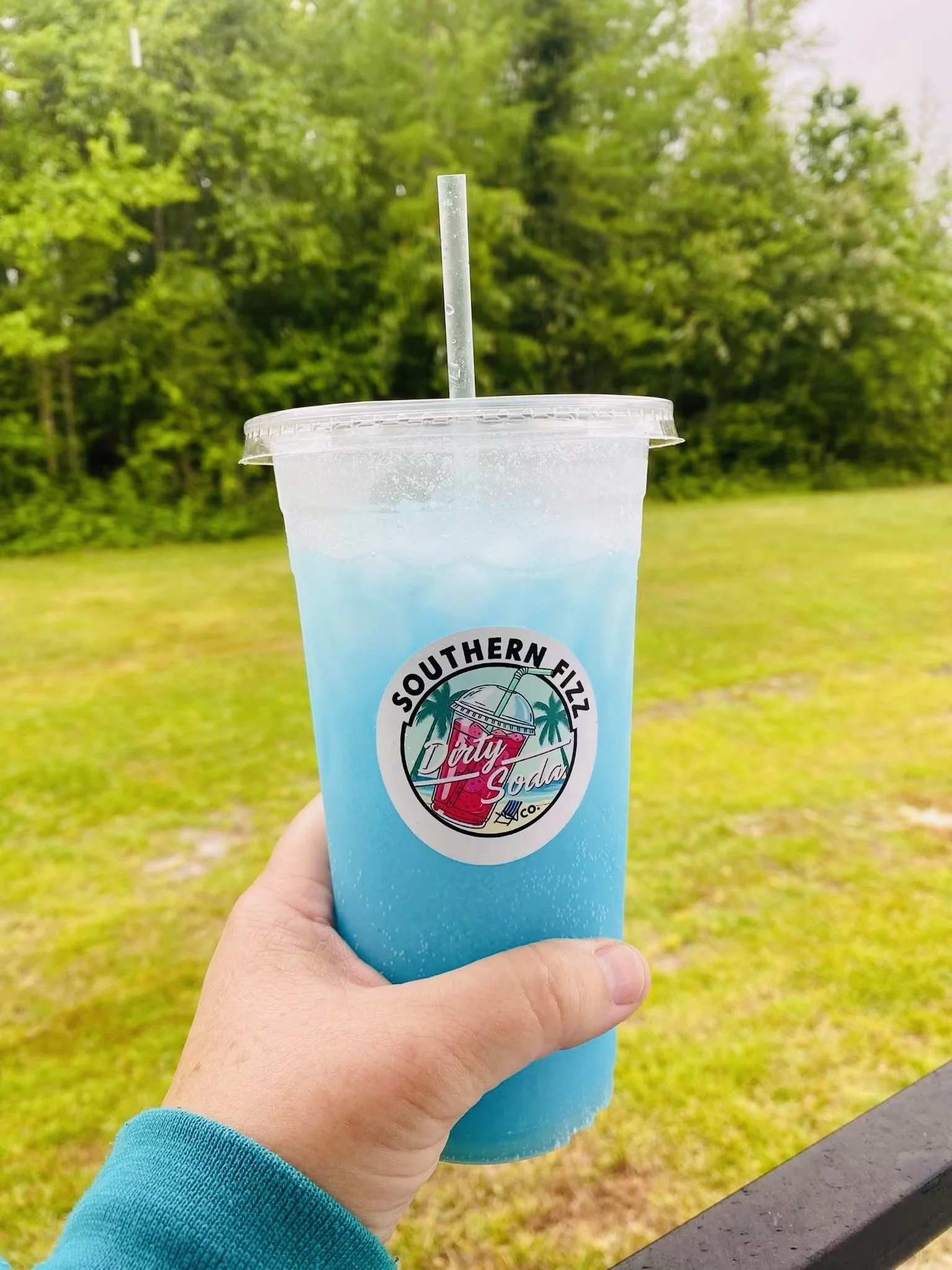 Hand holding a blue soda in a plastic cup with a straw, outdoors with green trees in the background.