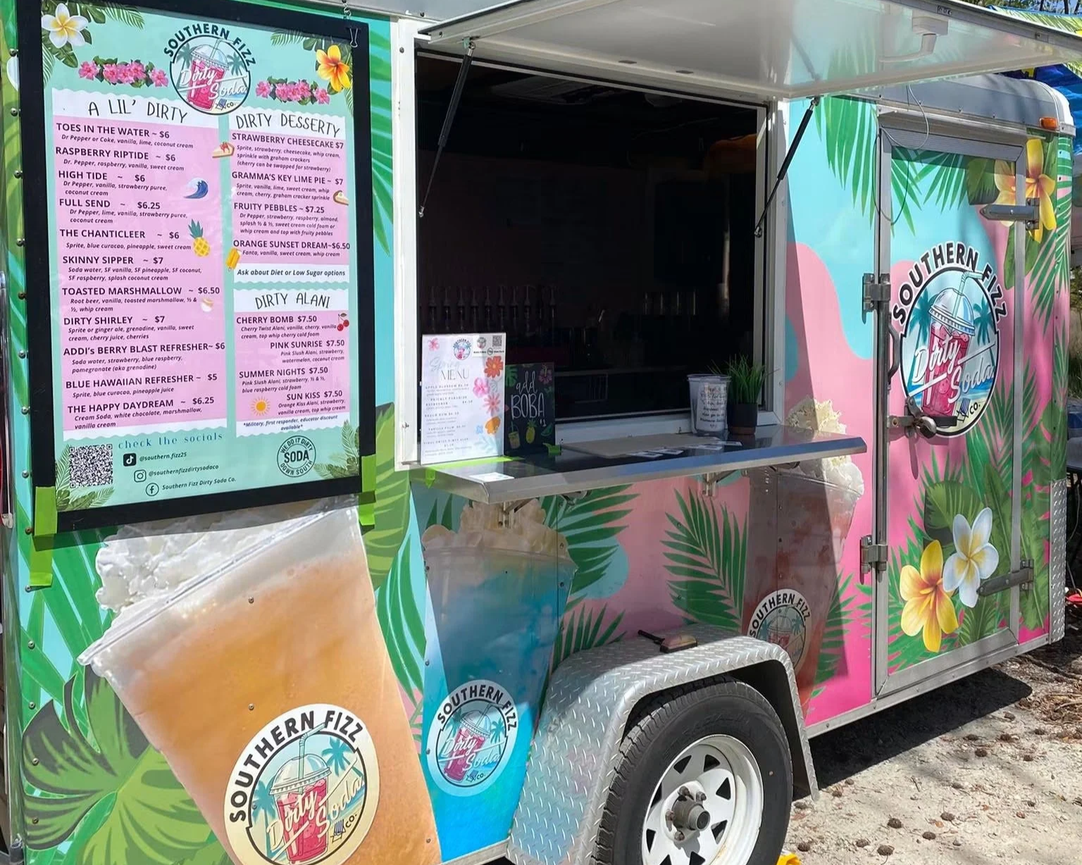 Colorful food truck with tropical floral and palm leaf decorations. The side menu lists various ice cream flavors and desserts. The truck has a large ice cream cone graphic and the logo 'Southern Fizz Dirty Soda Co.' on the side.