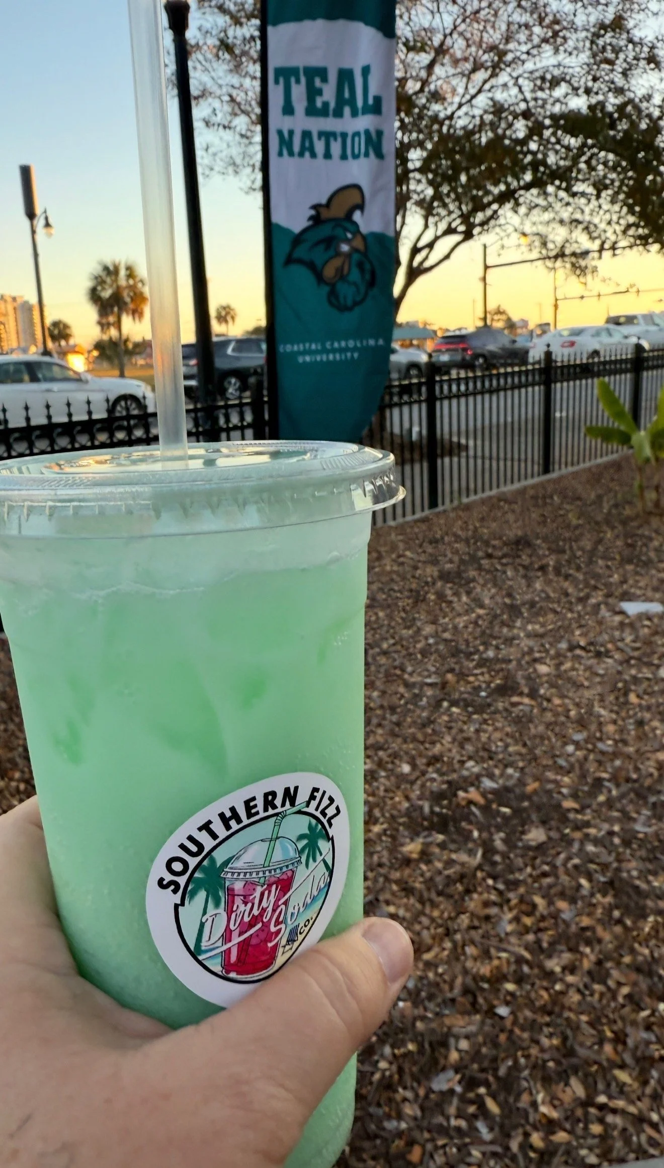 A hand holding a plastic cup with a green beverage and ice, with a logo that reads "SouthernFizz" and features a tropical drink. In the background, there is a parking lot, a banner for Coastal Carolina University with the text "Teal Nation" and a cartoon bird mascot, trees, and a sunset sky.