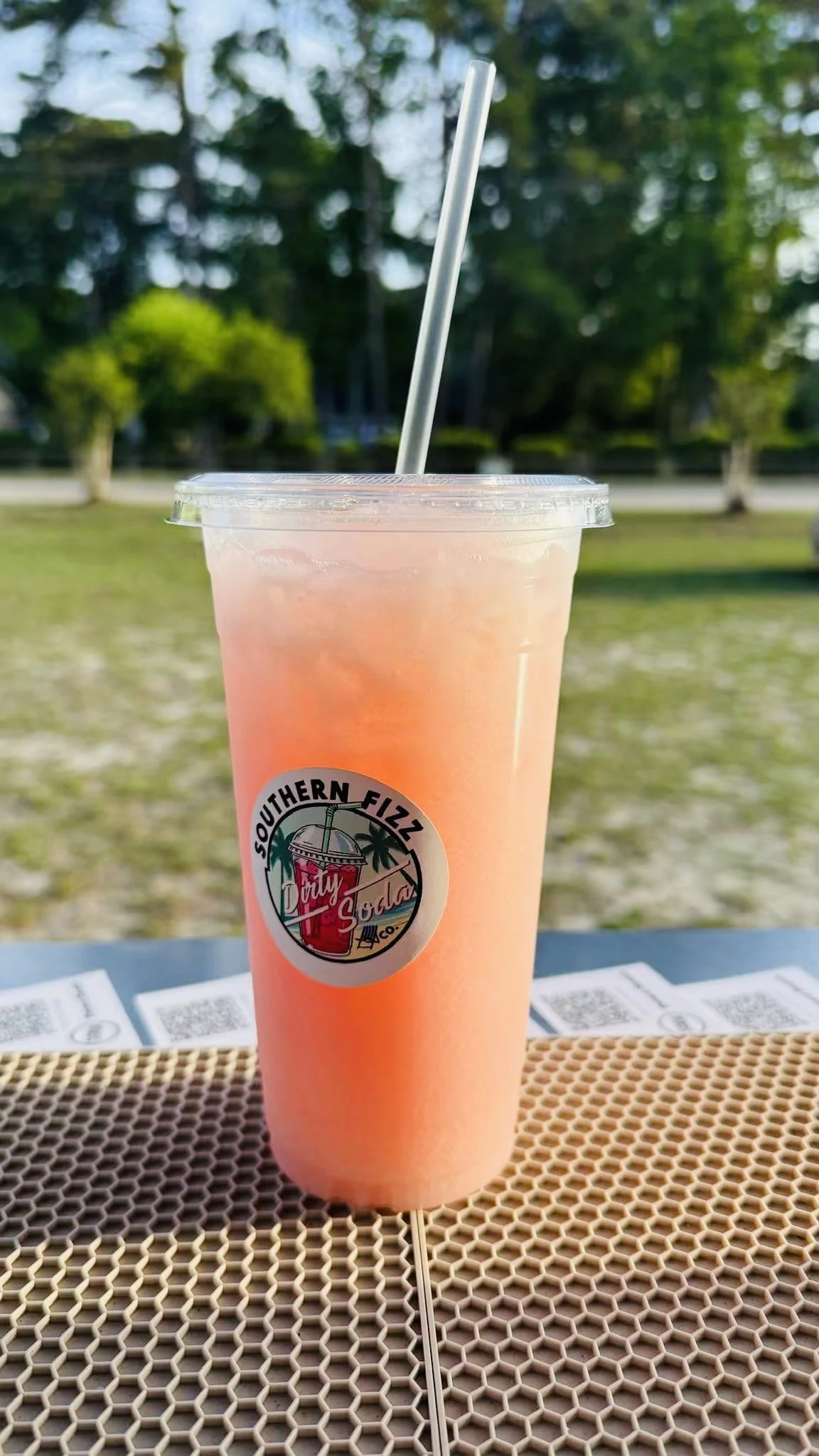 A pink frozen beverage in a clear plastic cup with a straw and a sticker that reads 'Southern Fizz' with a palm tree and a sunset. The cup is on a table outdoors with trees and a park in the background.