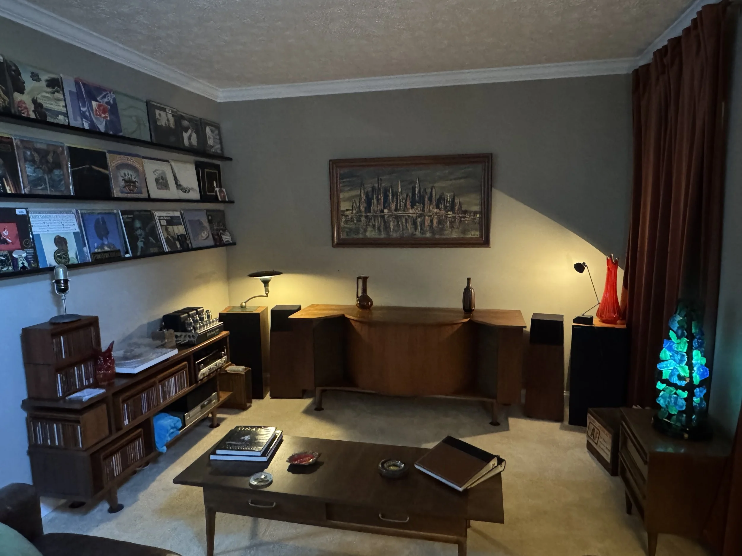 Living room with shelves of CDs and artwork, a wooden table with books and decorative items, a framed cityscape picture on the wall, a floor lamp, and a corner with a lamp and a colorful lamp/shade.