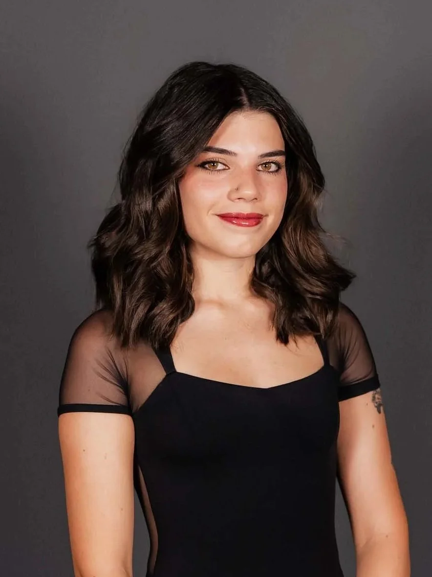 A woman with shoulder-length wavy dark brown hair, light skin, and makeup, wearing a black top with sheer fabric on the shoulders and smiling against a plain gray background.
