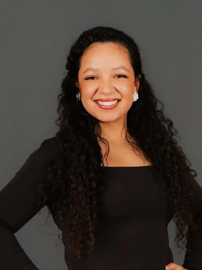 A woman with long, curly dark hair smiling and wearing a black top and pearl earrings against a dark gray background.