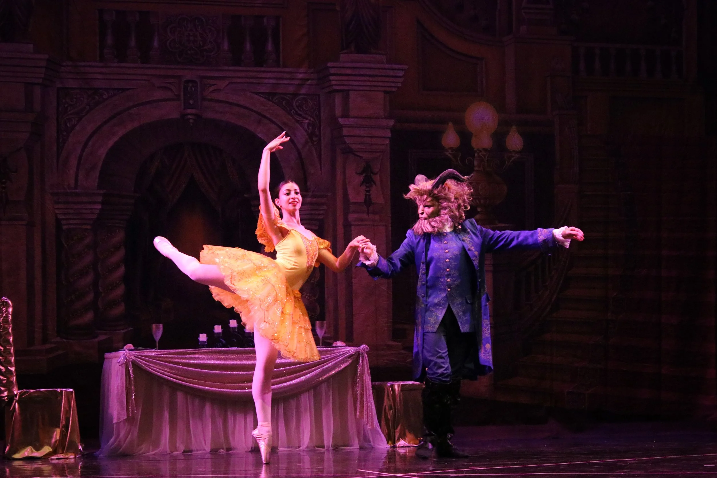 A ballerina in a yellow tutu dancing on stage with a man dressed as a beast. The stage has a dark wooden backdrop with ornate decorations.