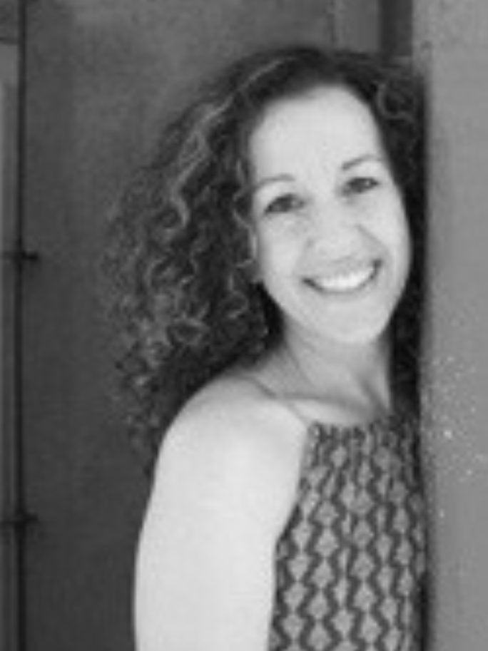 A woman with curly hair smiling and leaning against a wall in a sleeveless patterned top.